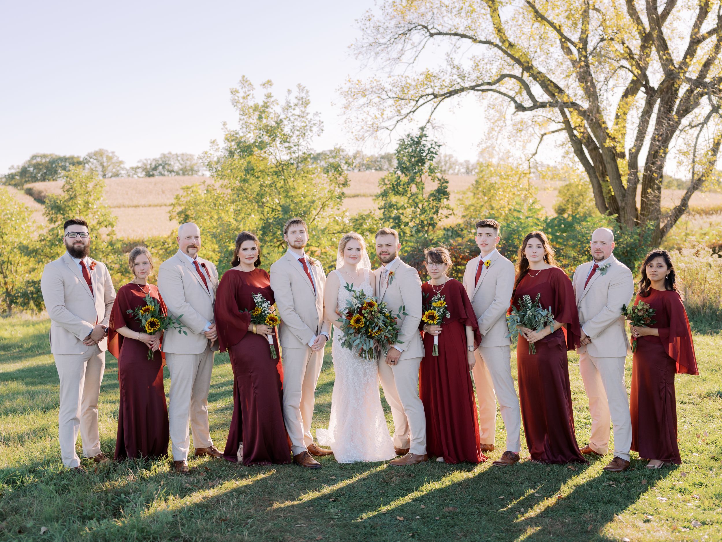Wedding party in burgundy and white attire poses together on a grassy hillside during golden hour.