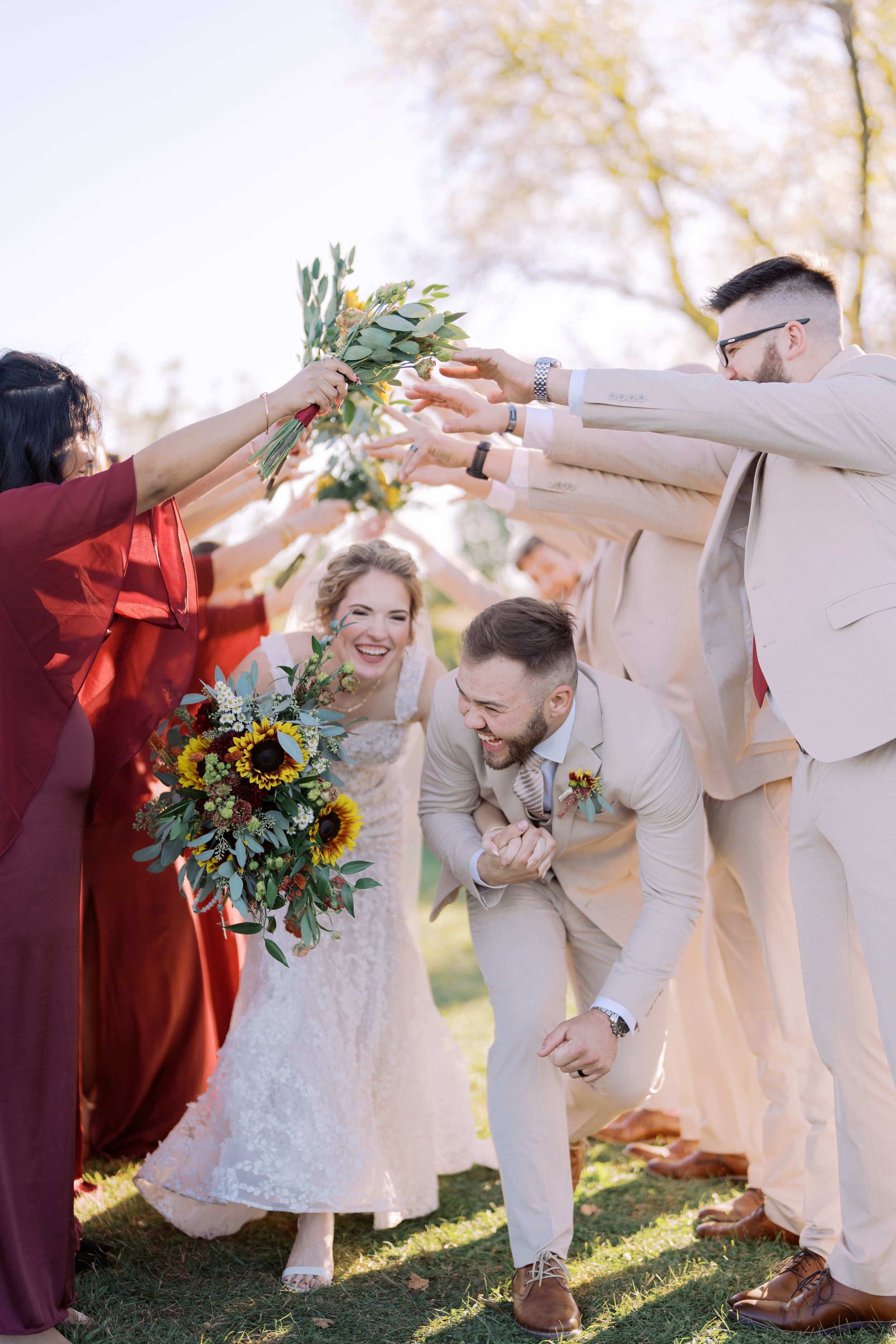 Wedding party celebrates joyfully on sunny lawn with sunflower bouquet and coordinated burgundy and beige attire.