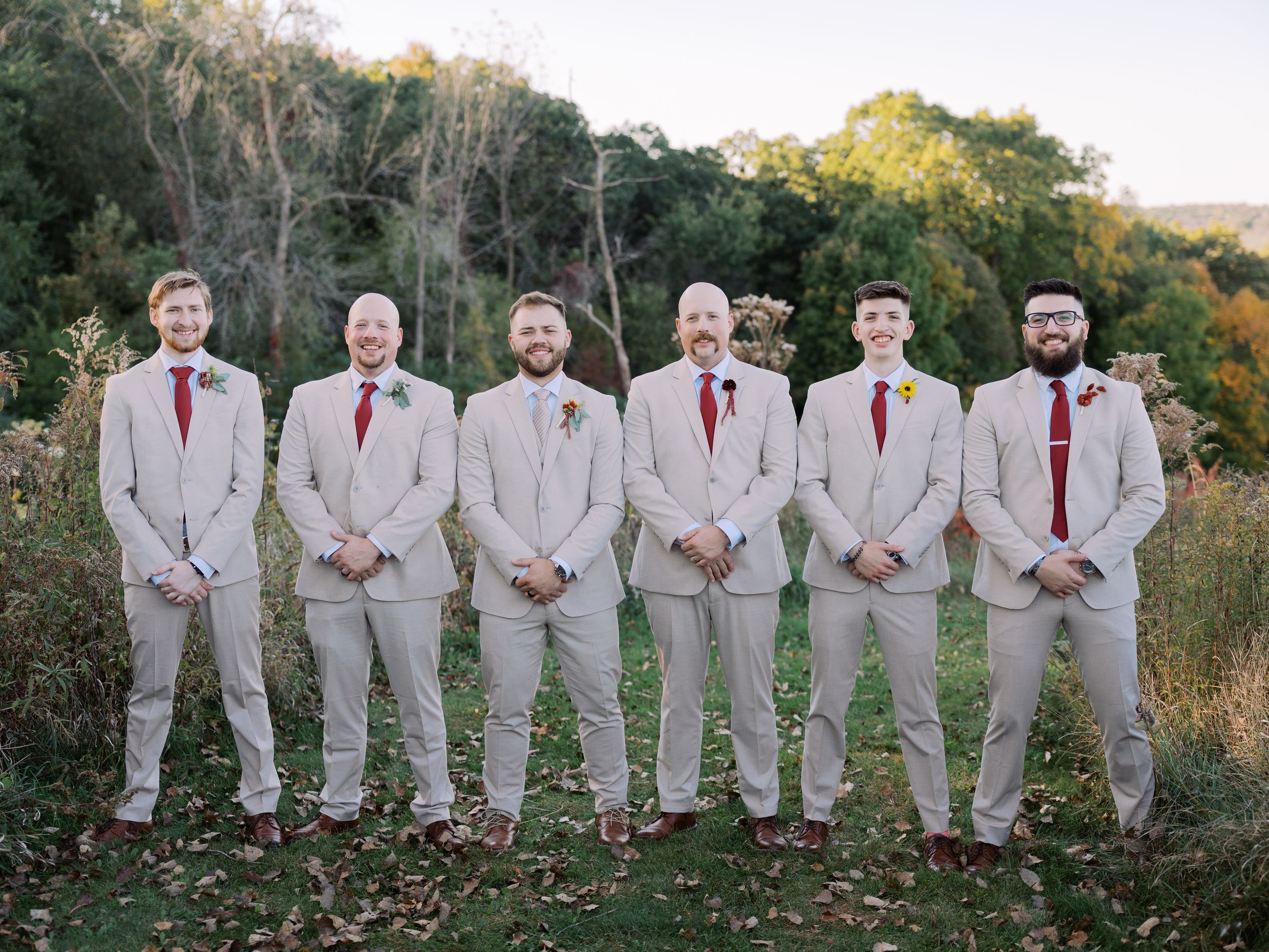 Wedding party wearing light gray suits with red ties pose together in an autumn outdoor setting.