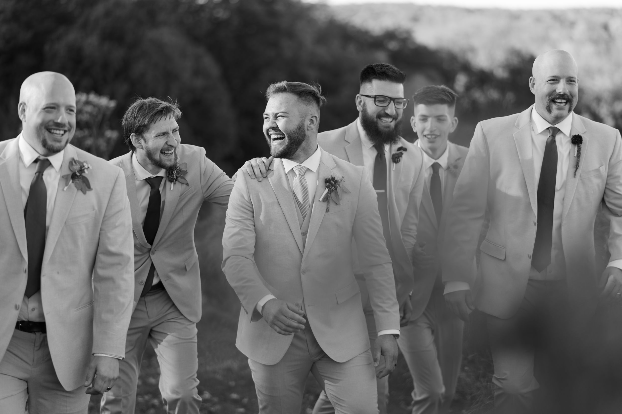 A group of groomsmen in light gray suits share a candid laughing moment together in black and white photography.