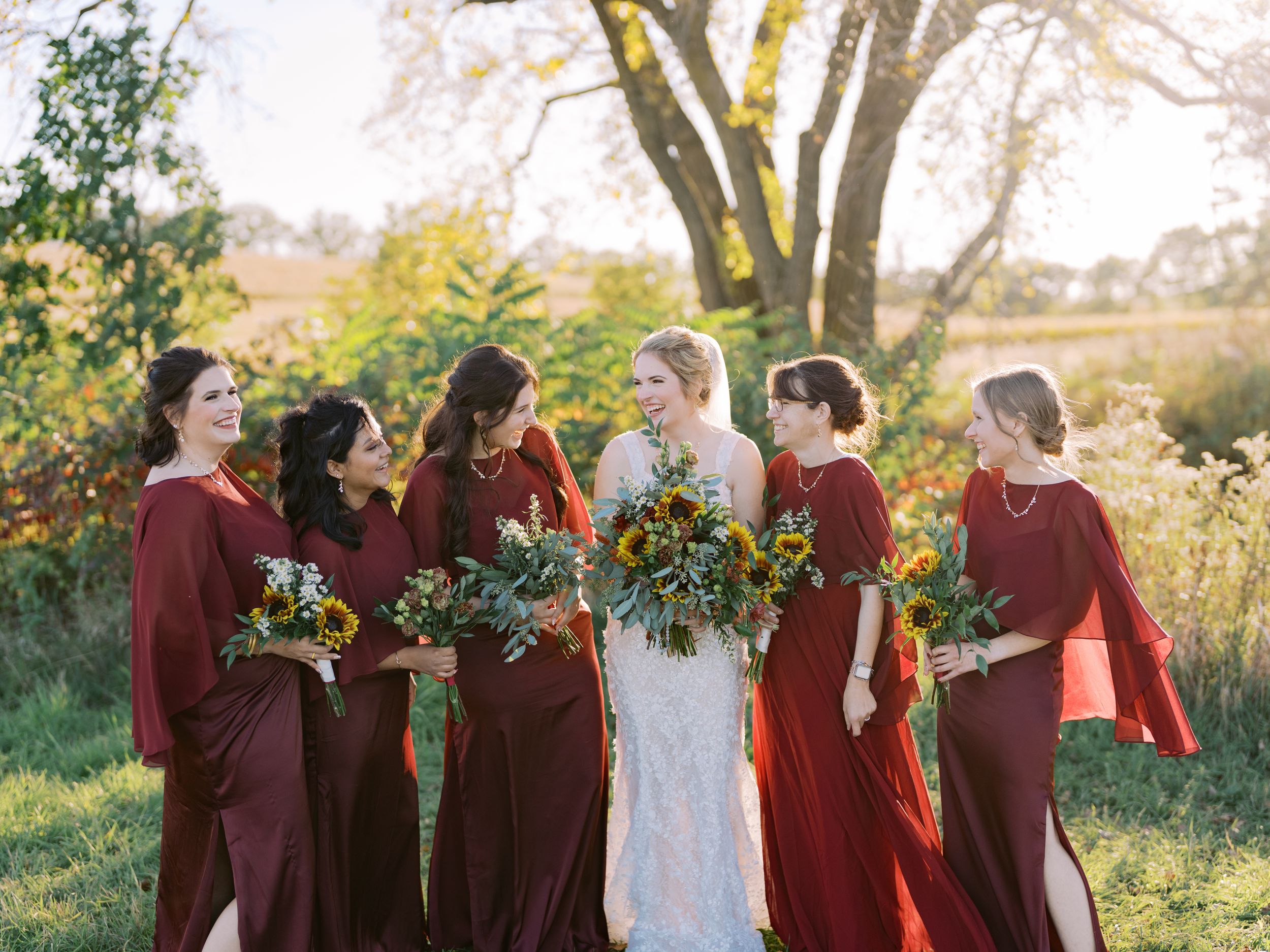 Wedding party in burgundy dresses holds vibrant fall bouquets while walking through a sunlit outdoor field.