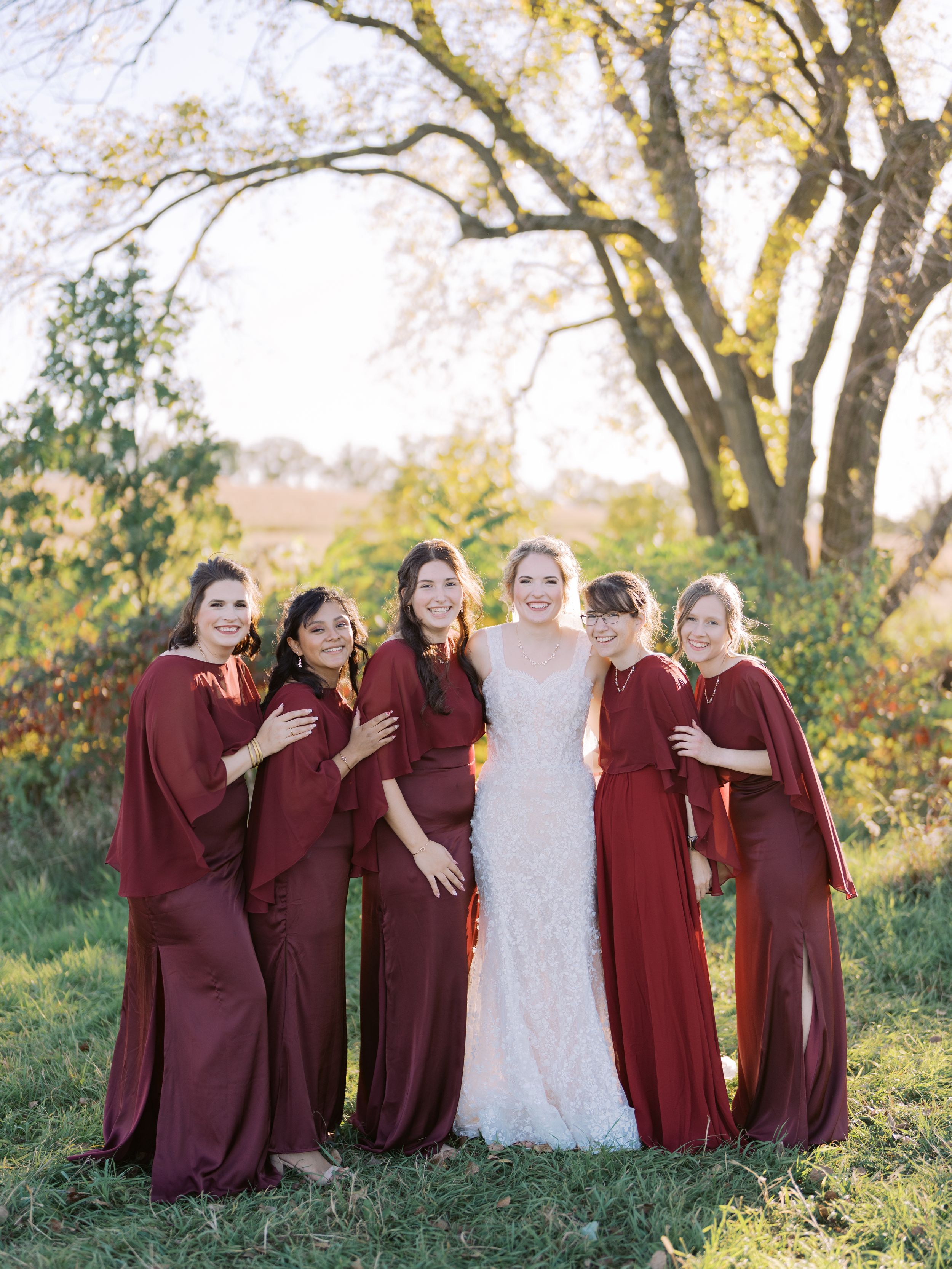 Bridal party in burgundy dresses and white gown pose together outdoors during autumn wedding photoshoot.