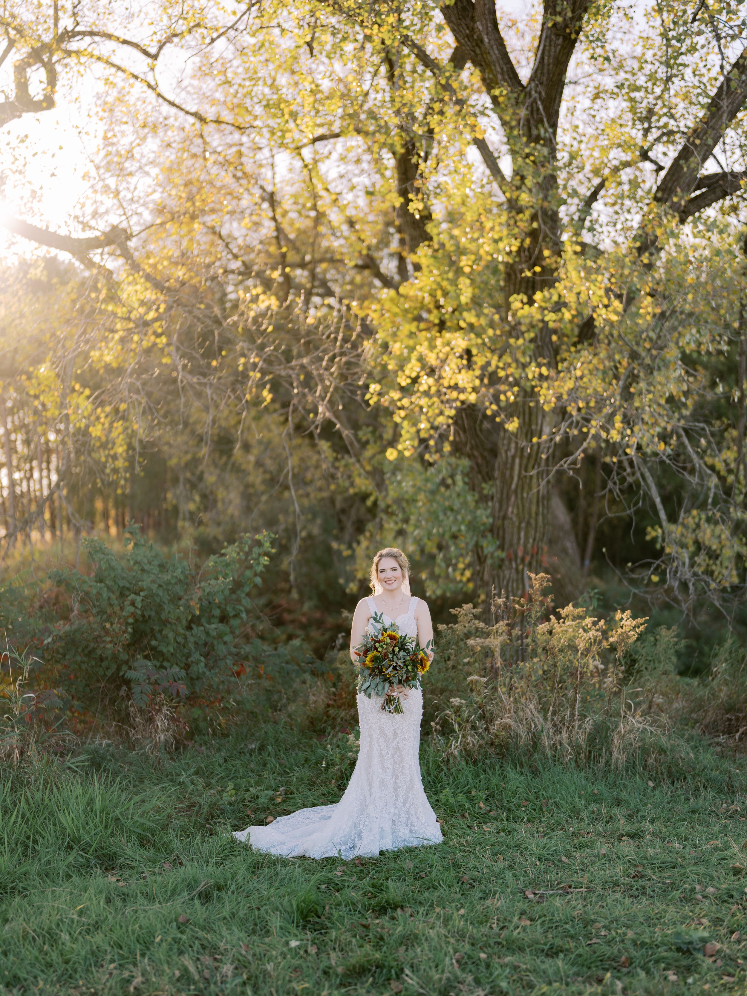 A figure in a flowing white wedding dress stands gracefully in a sunlit autumn forest setting.