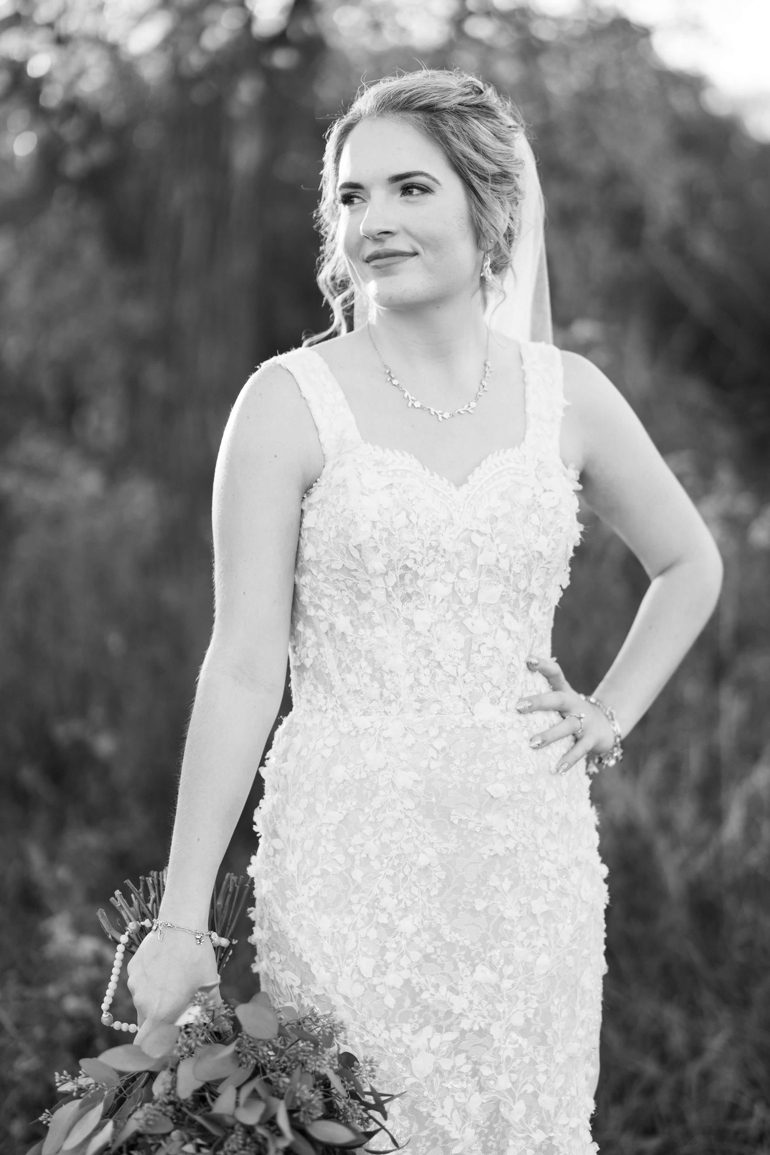 A bride in a detailed lace wedding dress poses outdoors holding a bouquet in black and white photography.