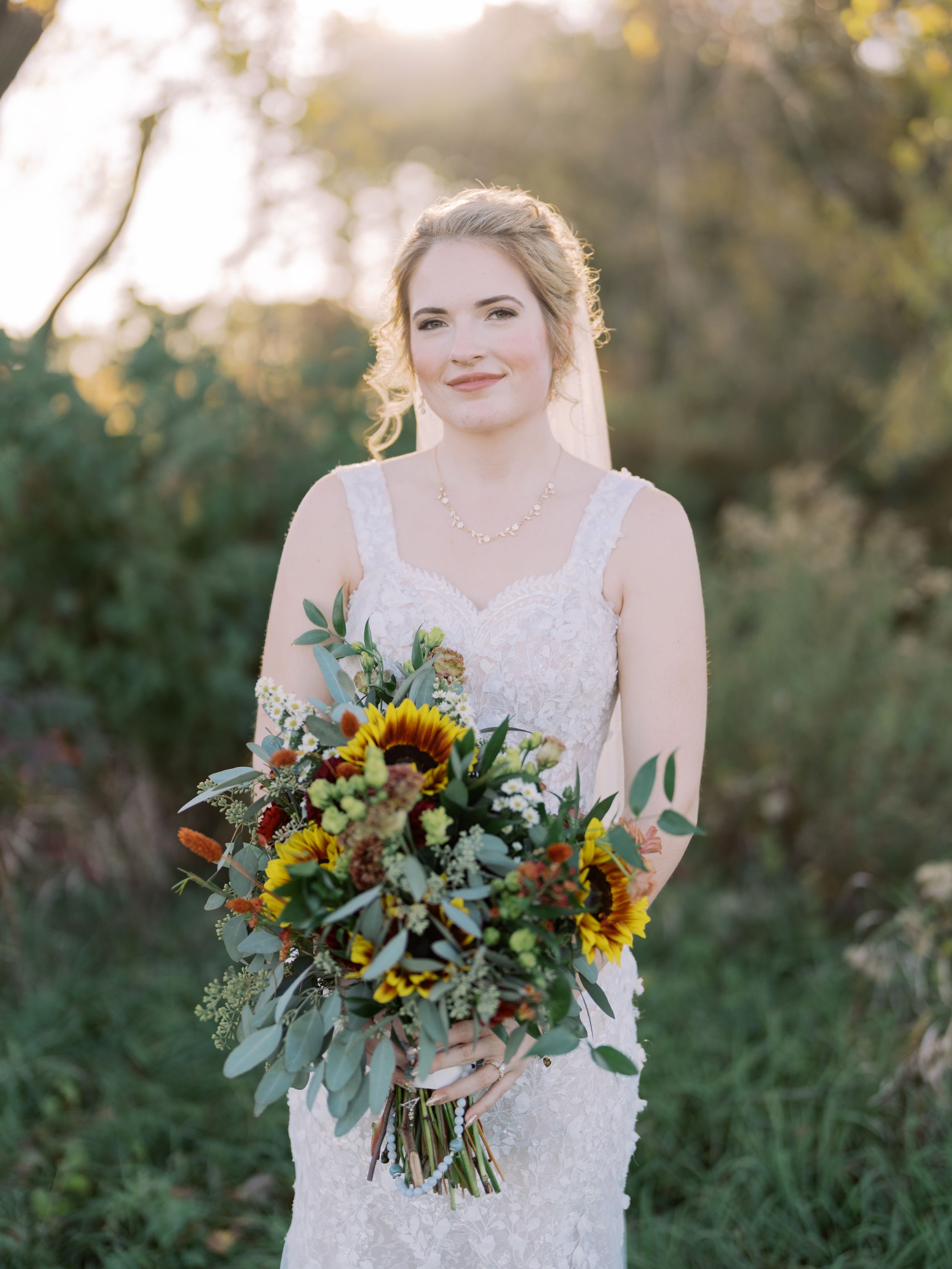 Bride in white lace dress holds vibrant sunflower and eucalyptus bouquet during outdoor sunset wedding photoshoot.