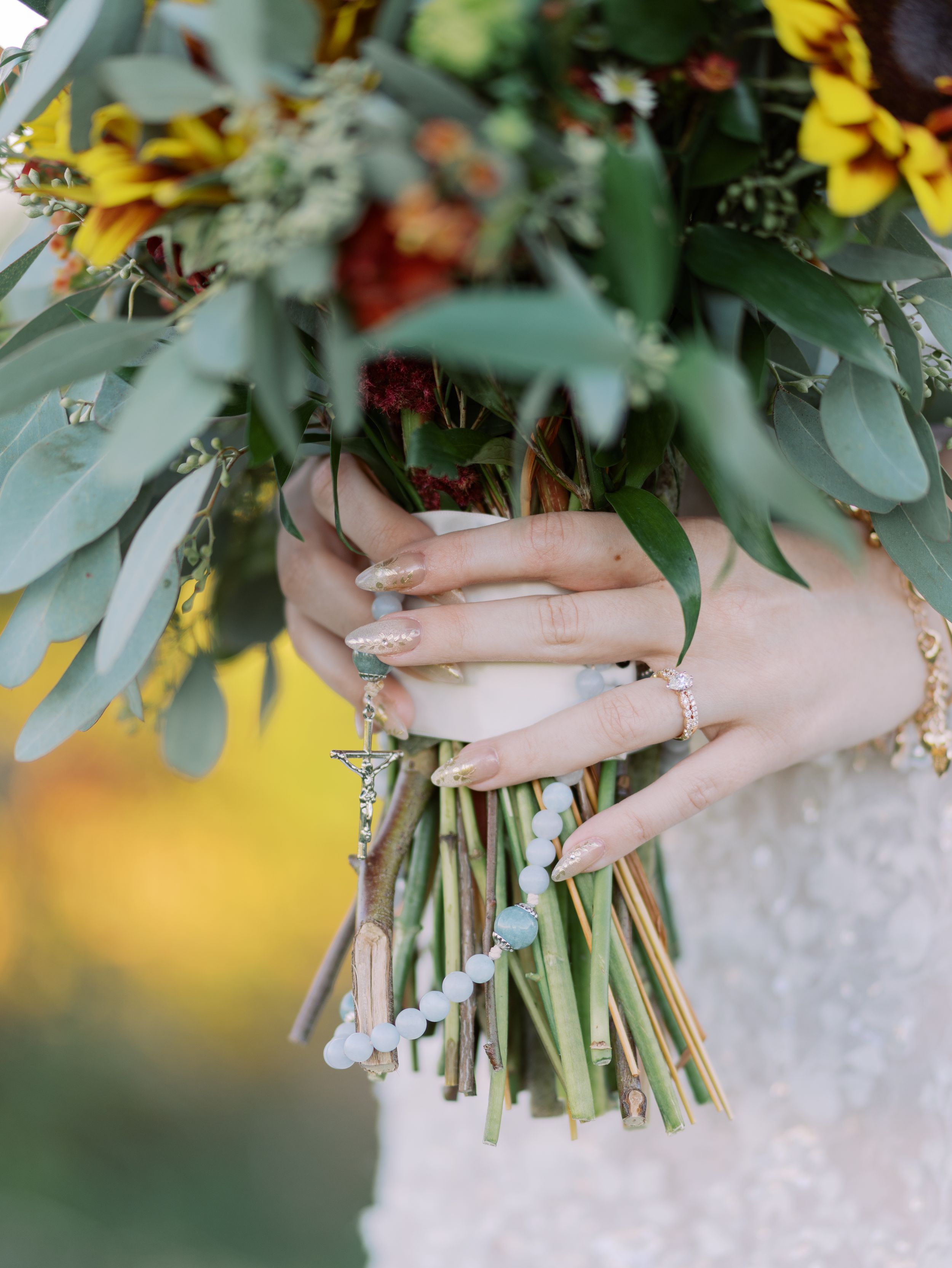 Hands holding a rustic wedding bouquet with yellow flowers and eucalyptus leaves, showcasing delicate rings and manicured nails.