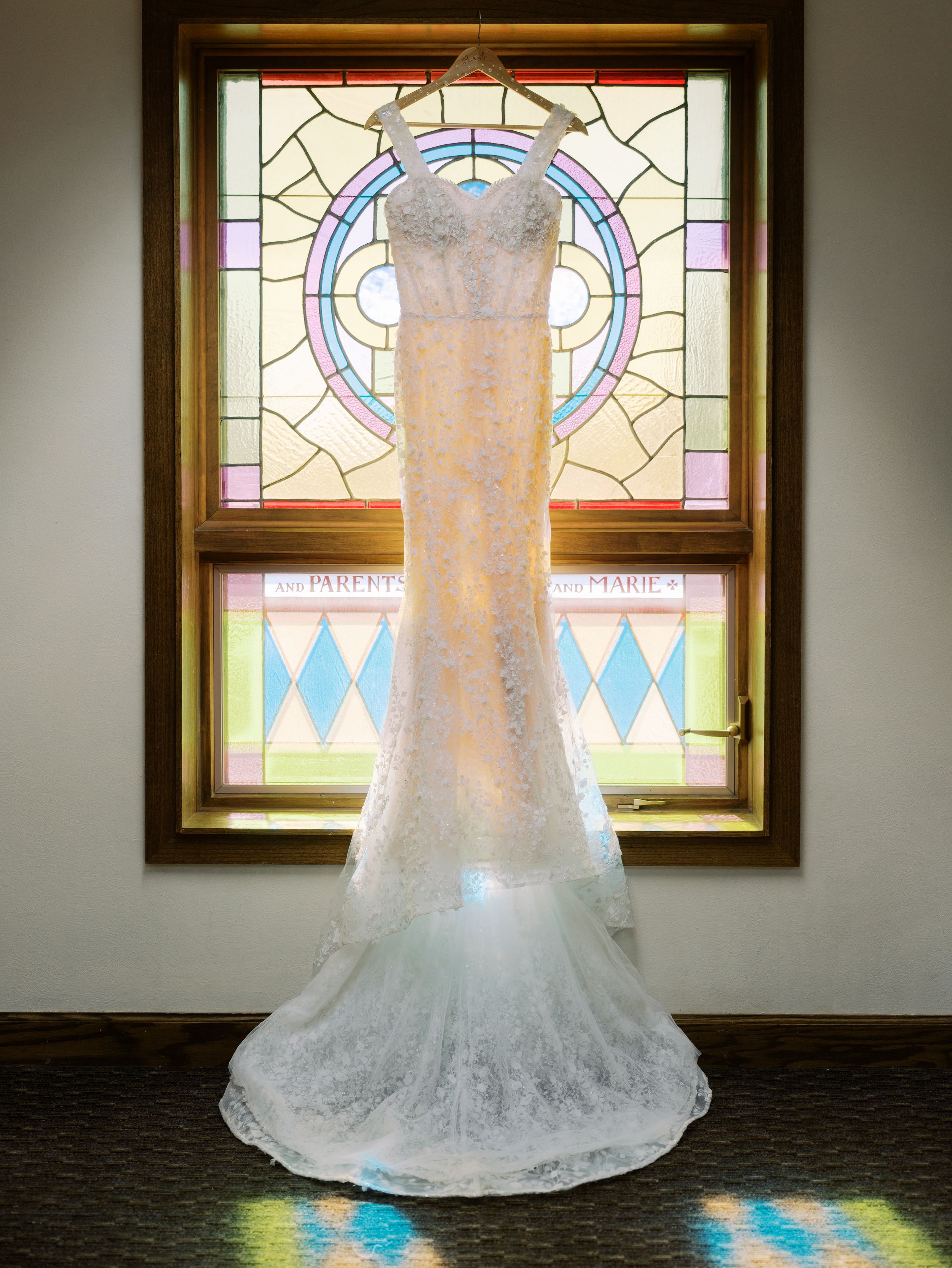 A white lace wedding dress is displayed in front of a stained glass window creating a beautiful backlit effect.