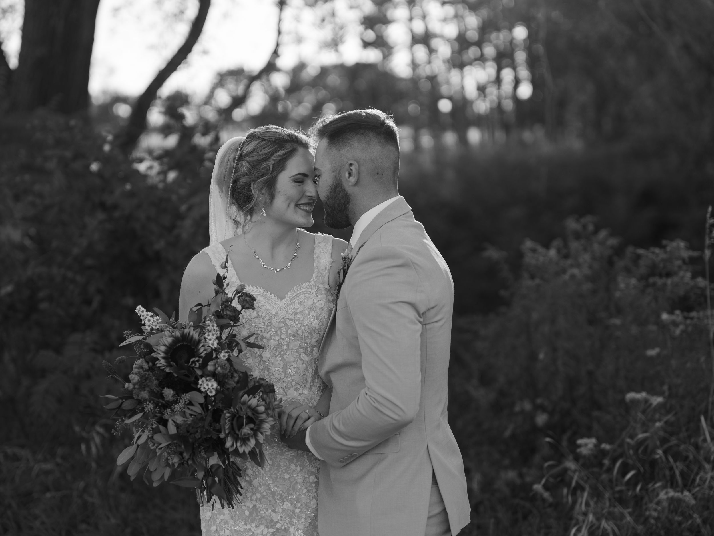 A couple embraces during an outdoor wedding ceremony in a garden with lush greenery and trees in the background.