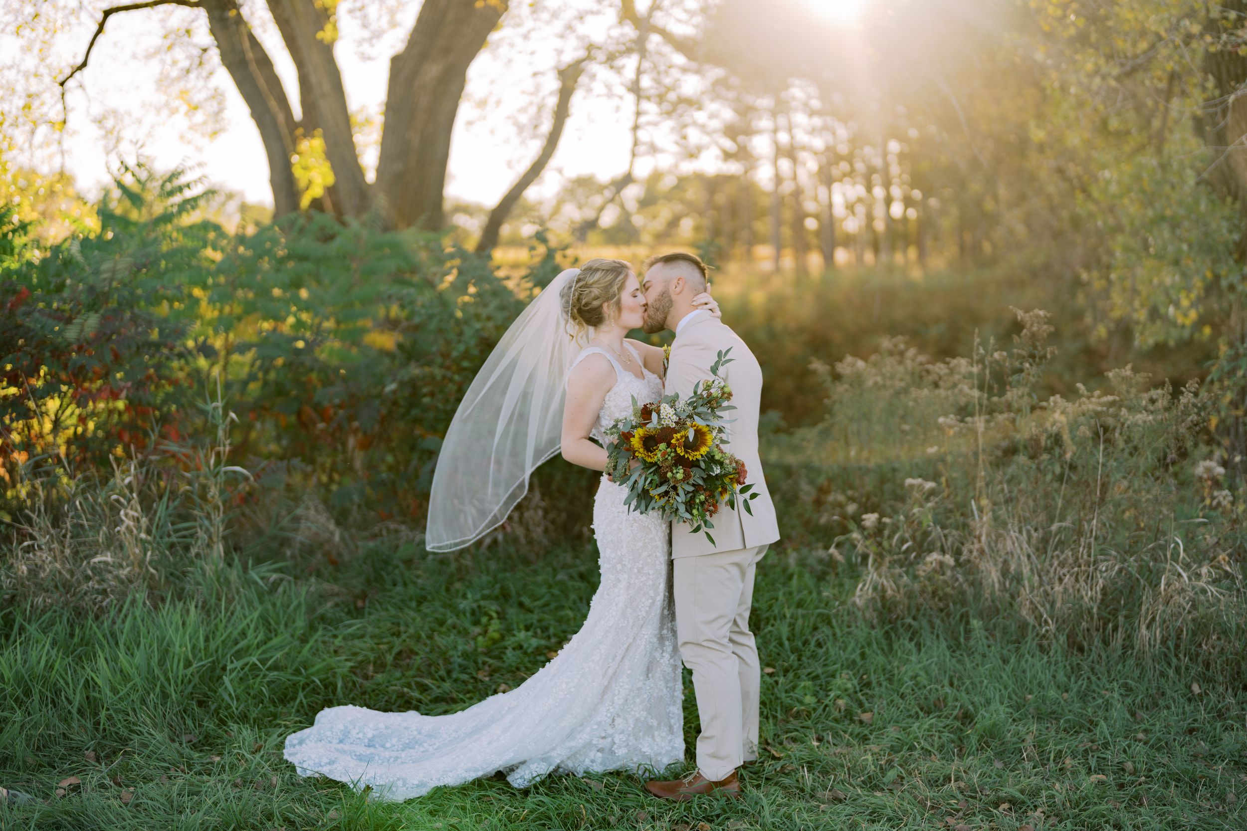 A romantic wedding couple embraces in a sunlit meadow during golden hour with flowing white dress and veil.