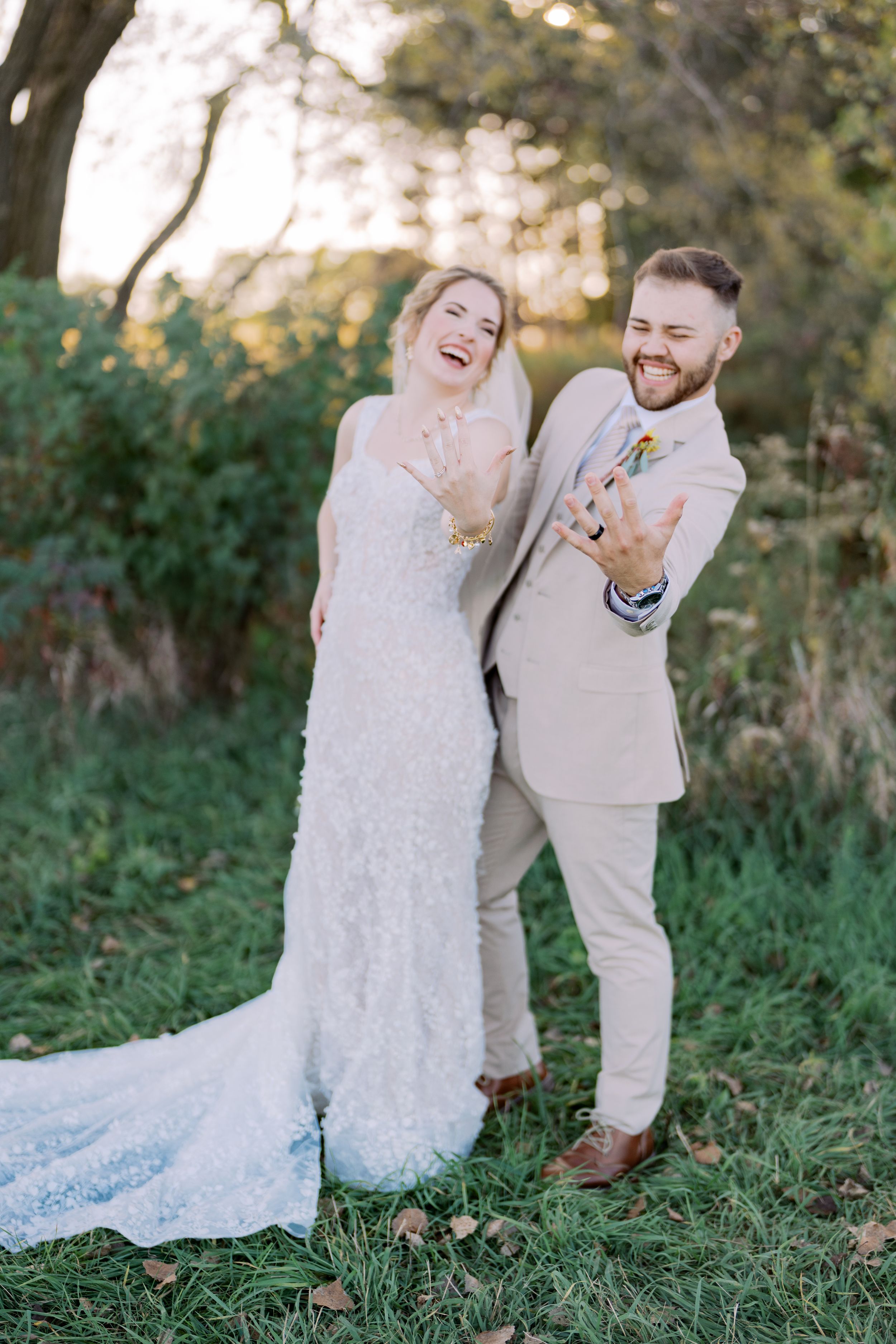 Newlywed couple laughs together while sitting in a grassy meadow during golden hour sunset.