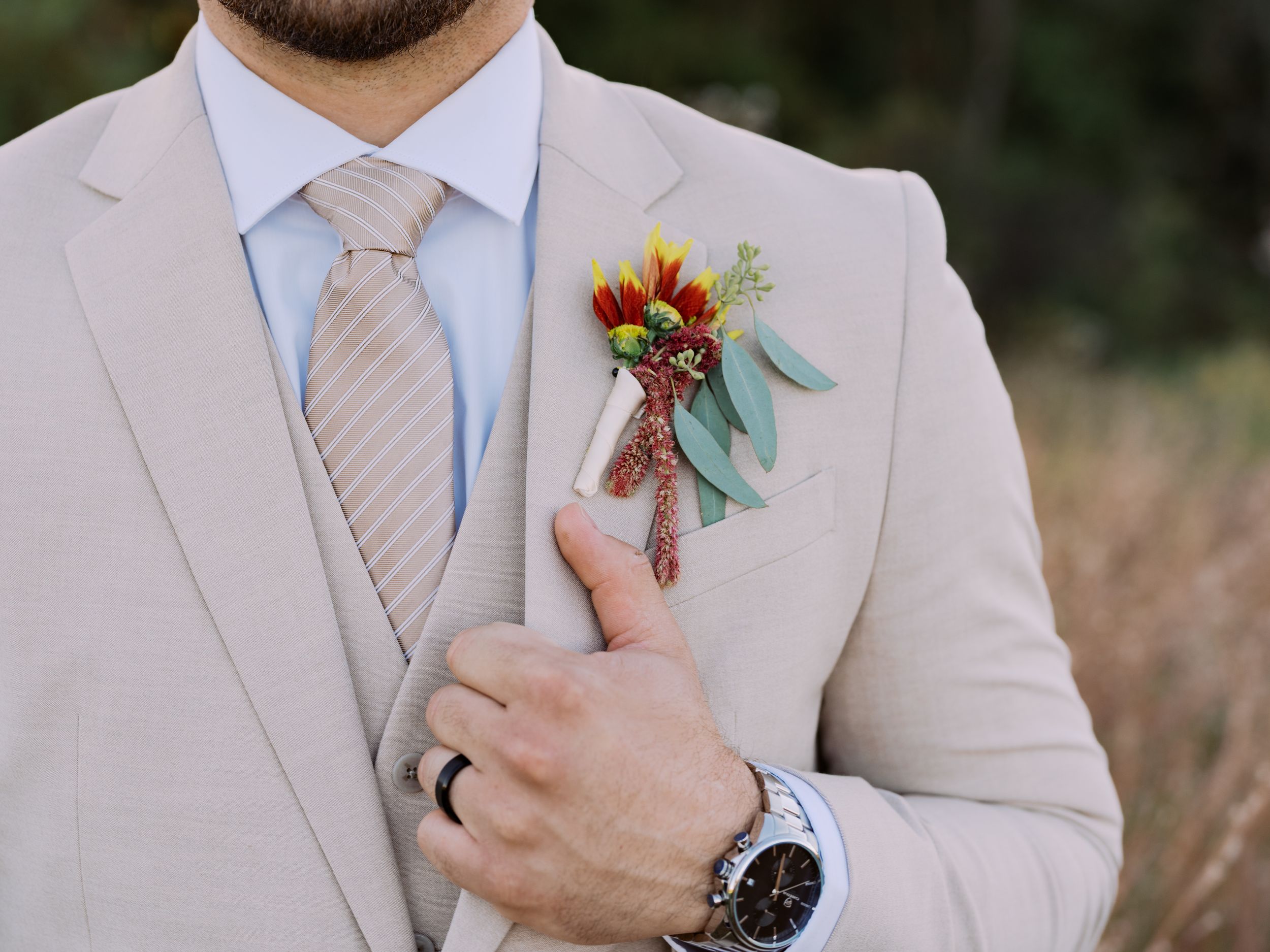 Close up of a beige suit jacket with striped tie and colorful fall boutonniere featuring sunflowers and teal ribbon.