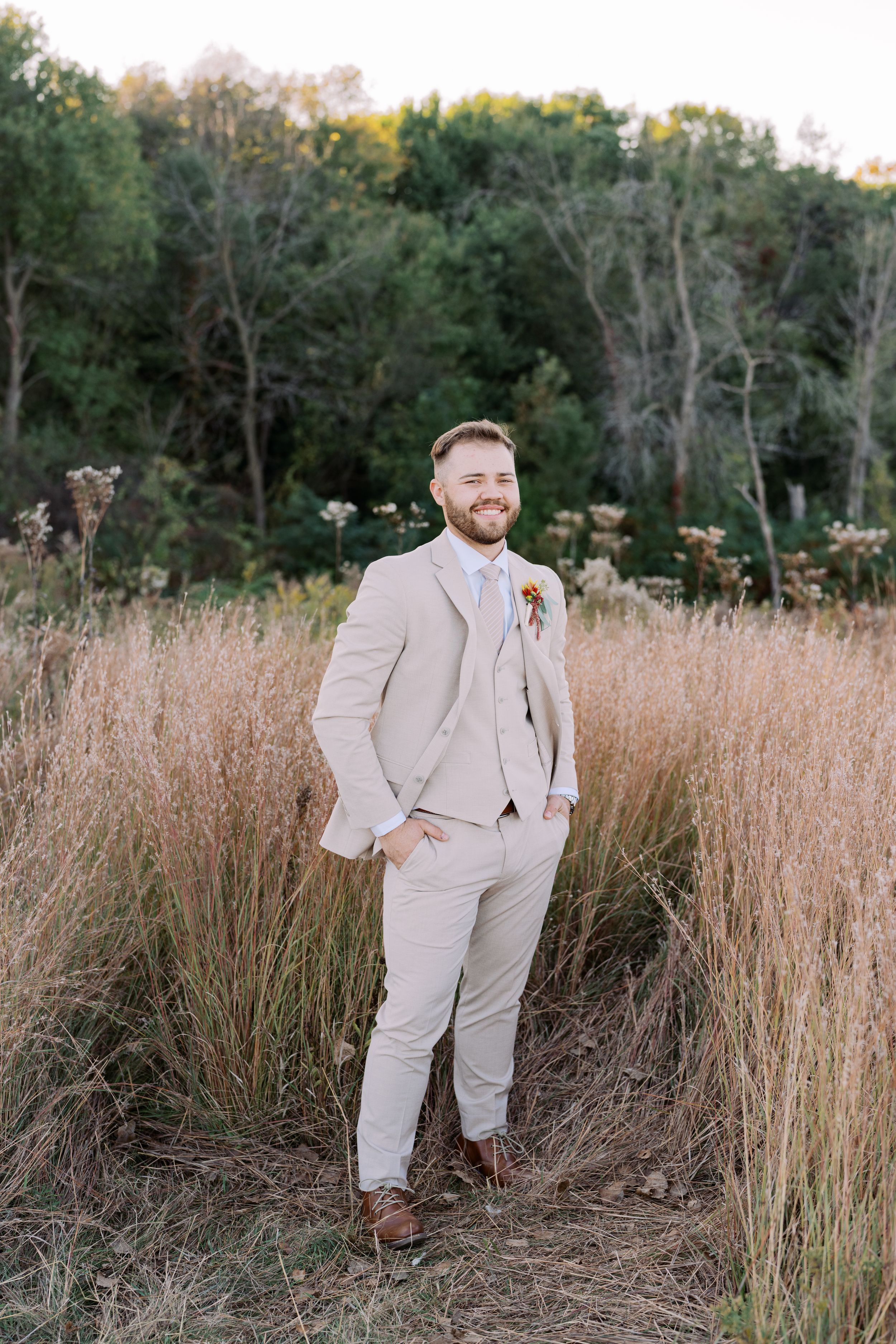 A person in a light beige suit stands in a field of tall grass during golden hour with trees in the background.