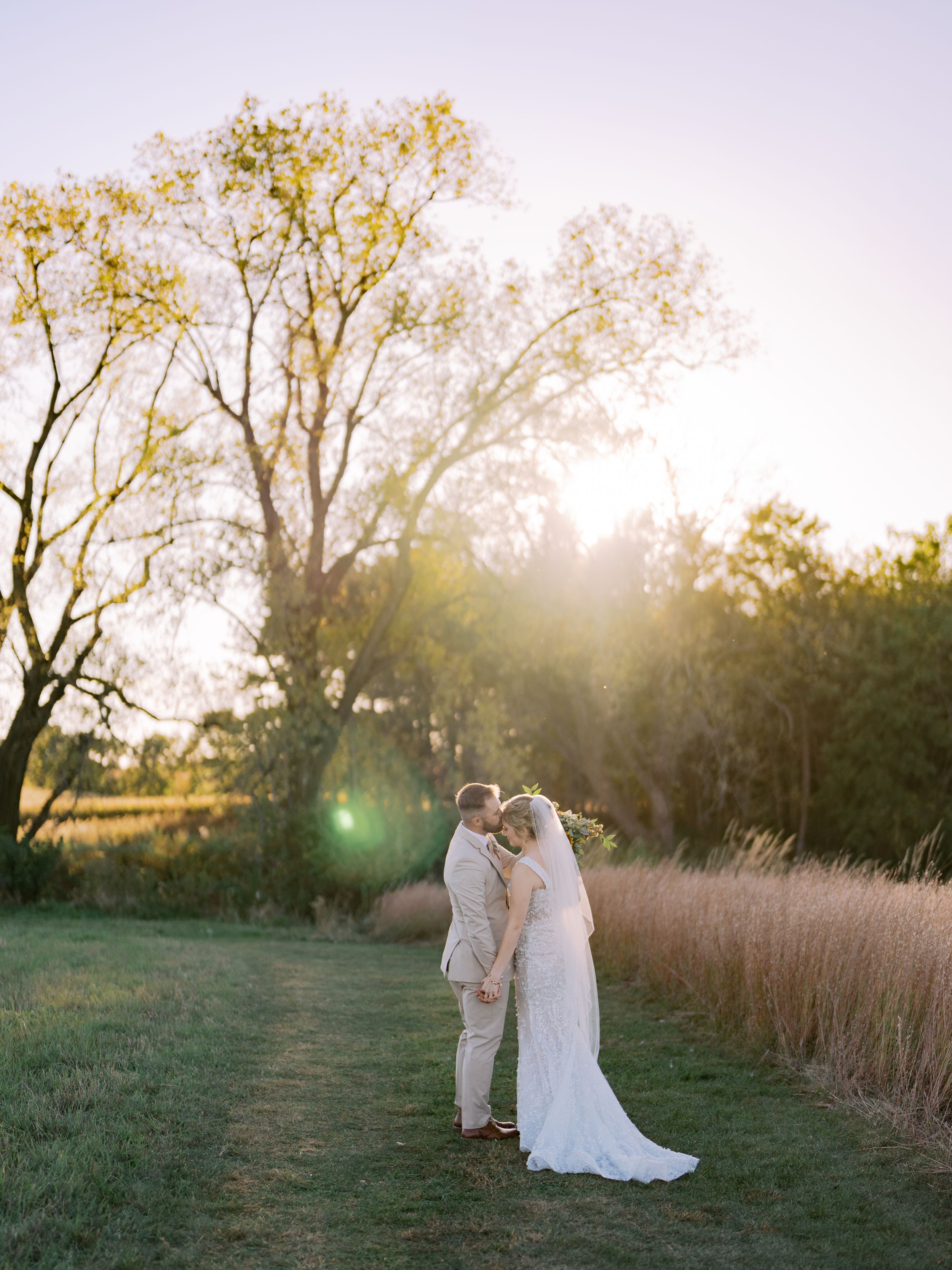 A romantic couple embraces during golden hour in a rustic field with tall grass and trees.