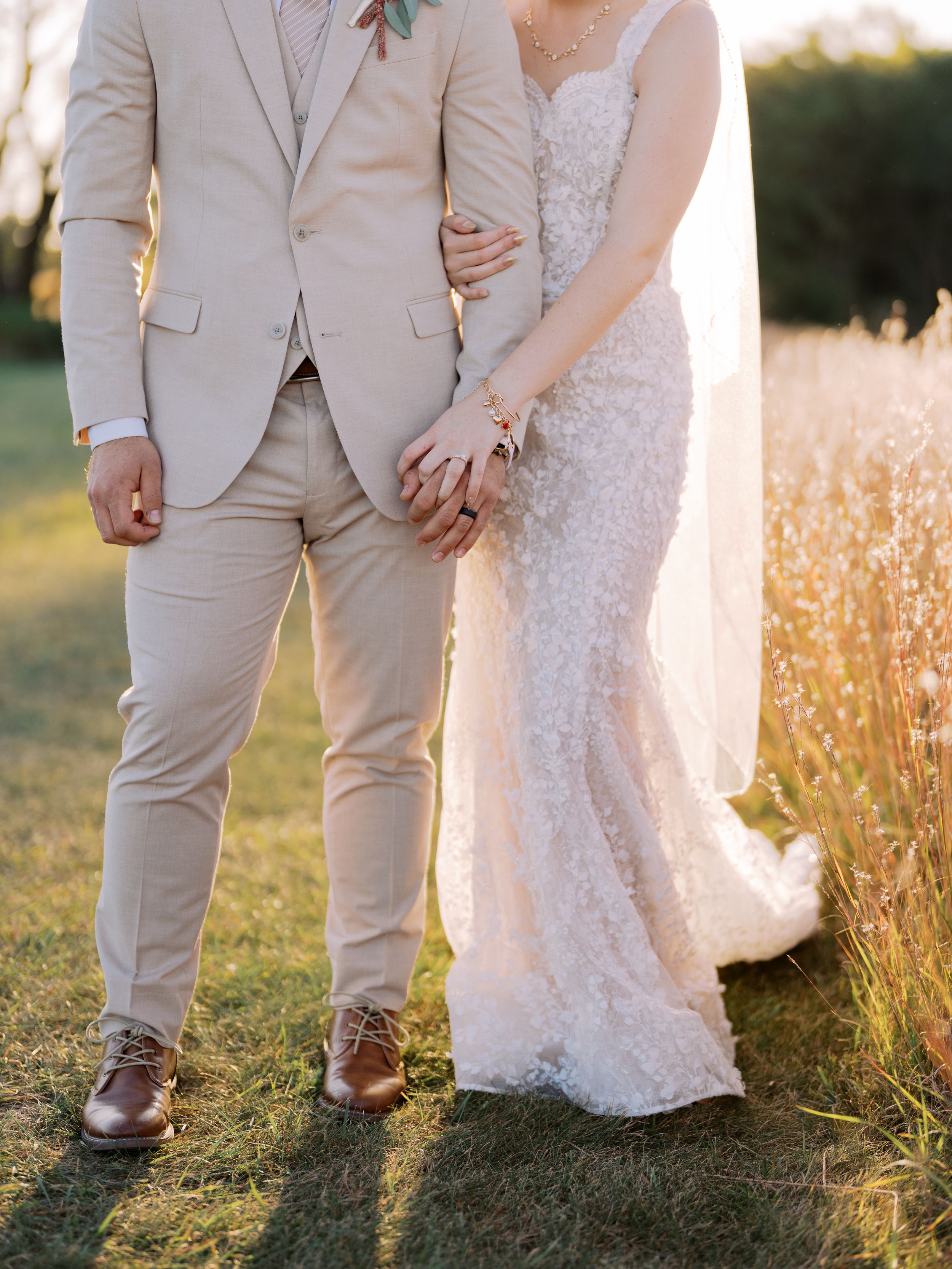 A romantic wedding couple stands in a golden wheat field at sunset wearing white dress and beige suit.