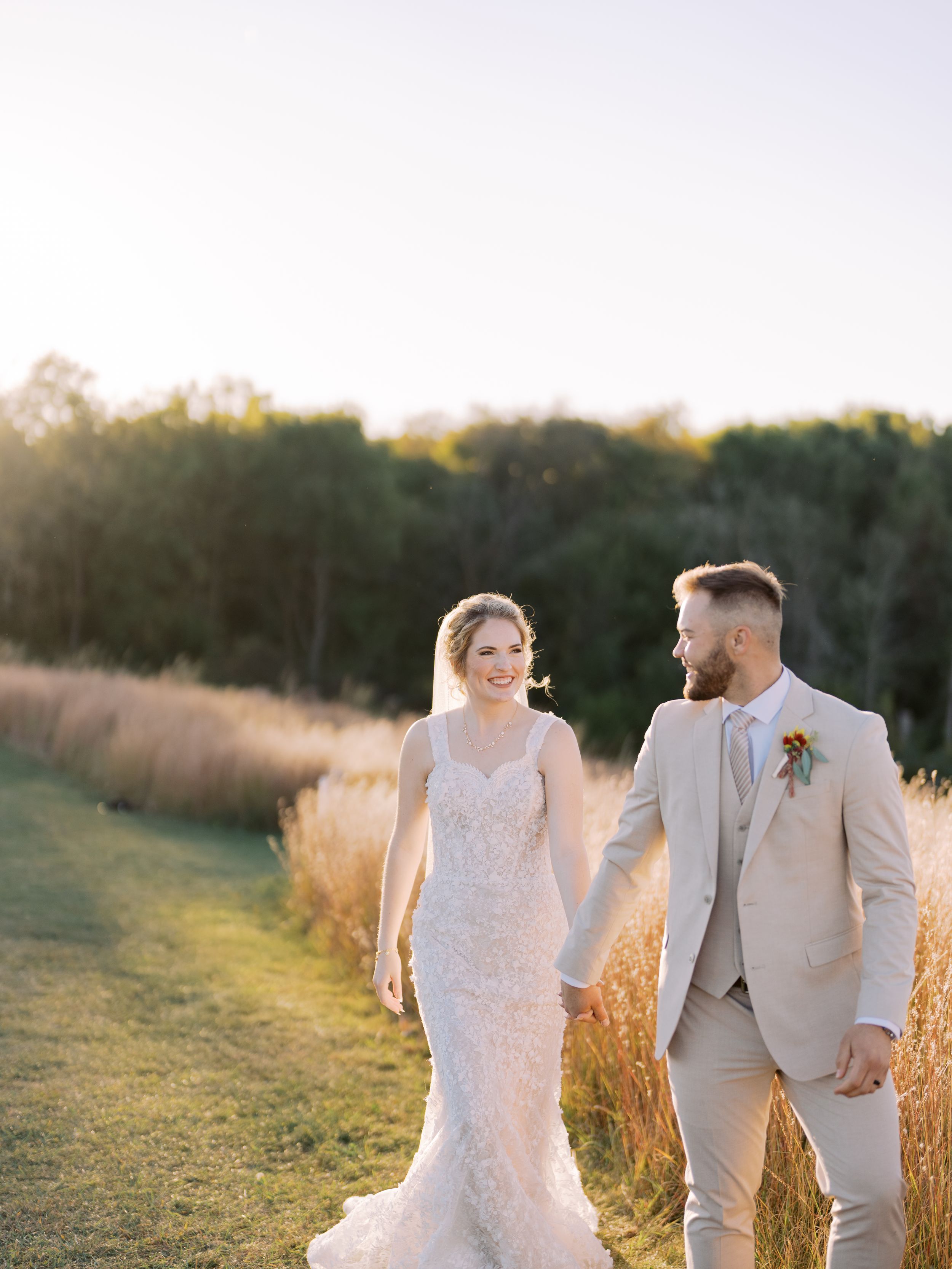 A bride and groom walk hand in hand through a golden field at sunset wearing beige and white wedding attire.