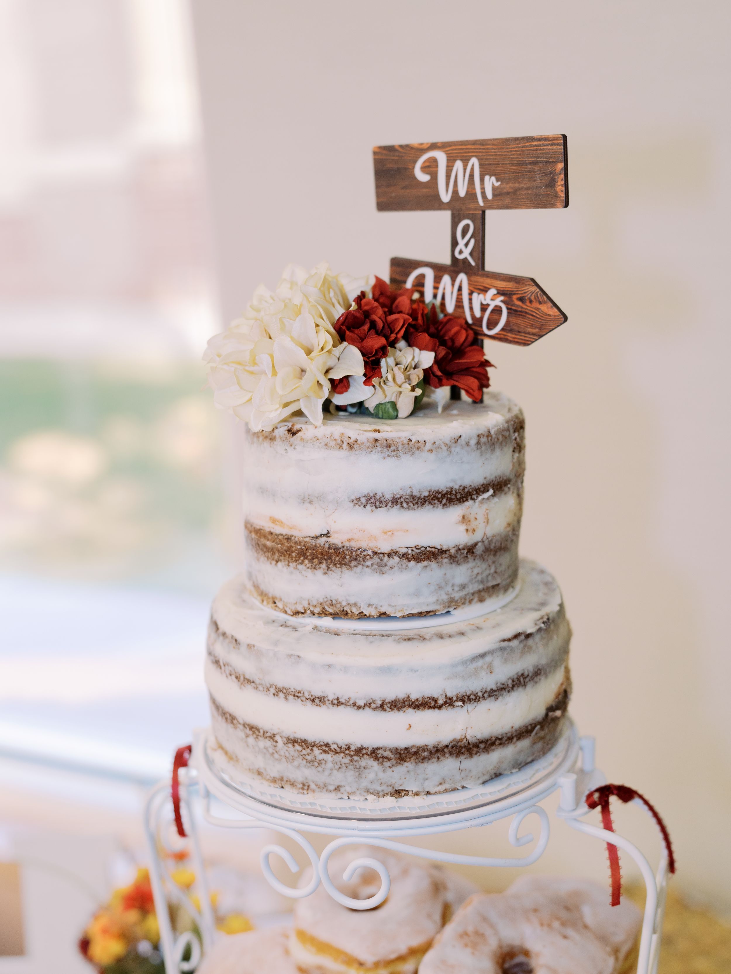 A rustic semi-naked wedding cake with wooden Mr and Mrs toppers decorated with burgundy flowers and greenery.