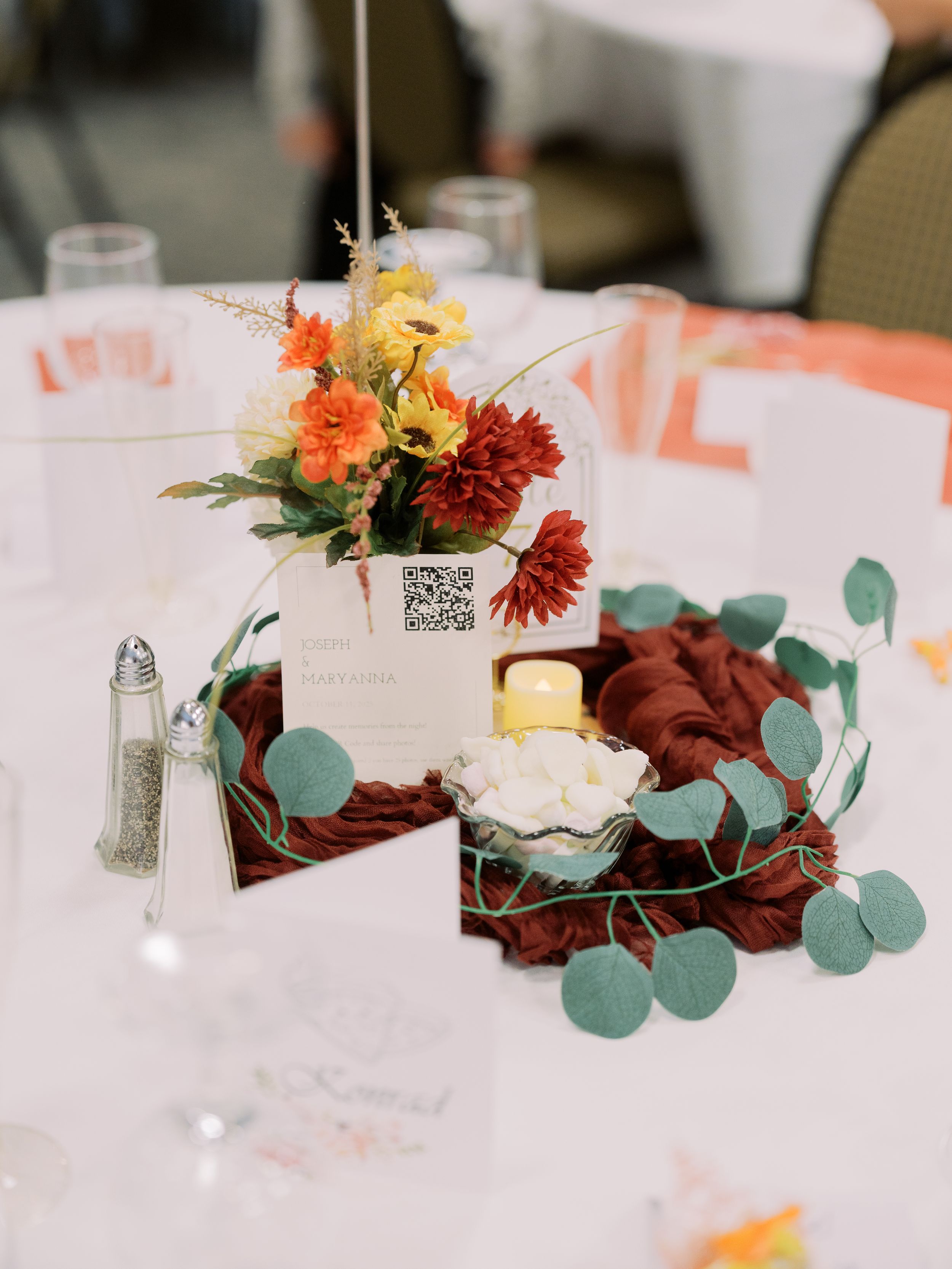 Wedding table centerpiece with orange and red flowers in a white vase, surrounded by eucalyptus leaves on burgundy fabric.