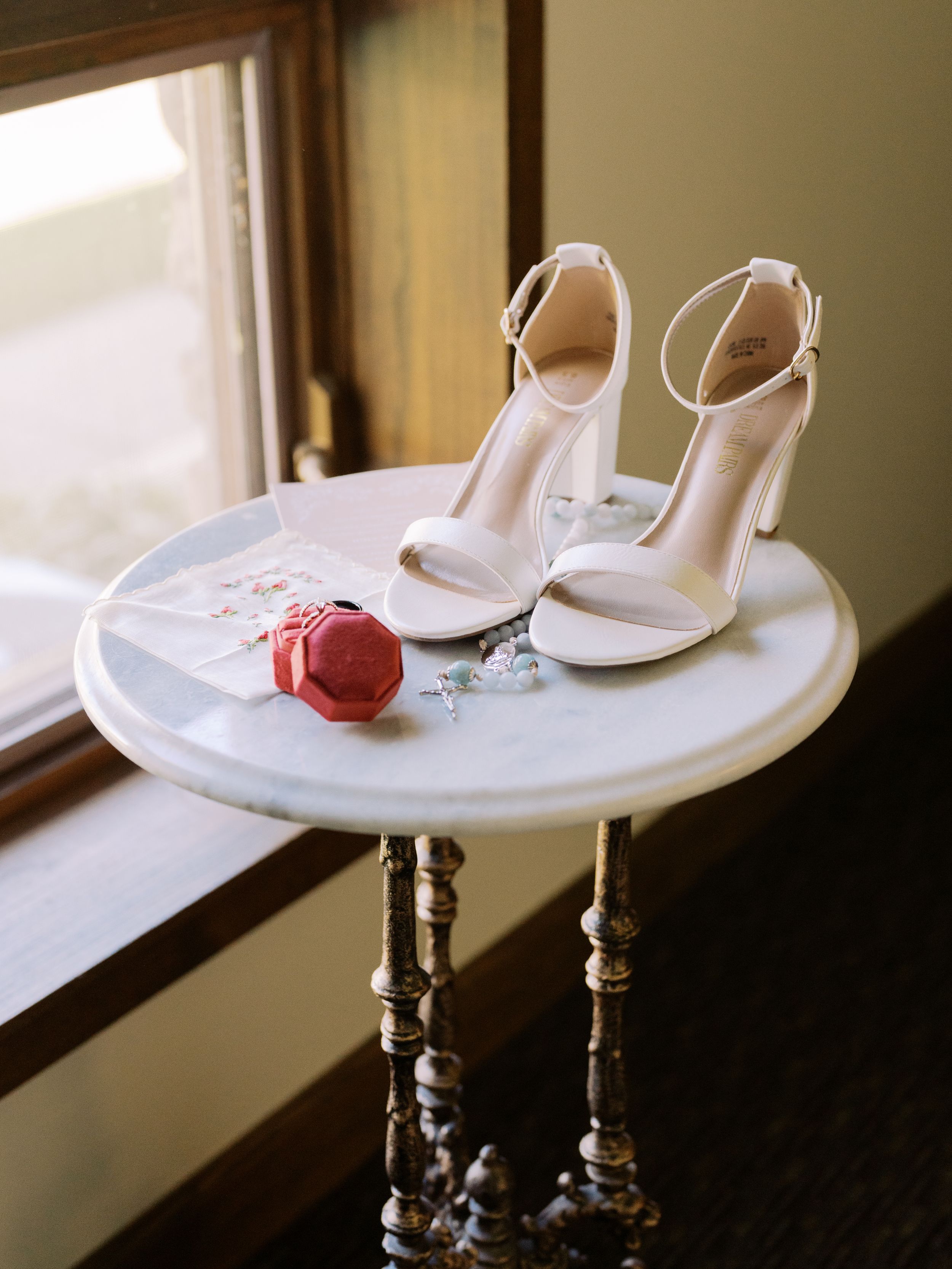 White bridal shoes and a pink macaron sit elegantly on a round marble table by a window.