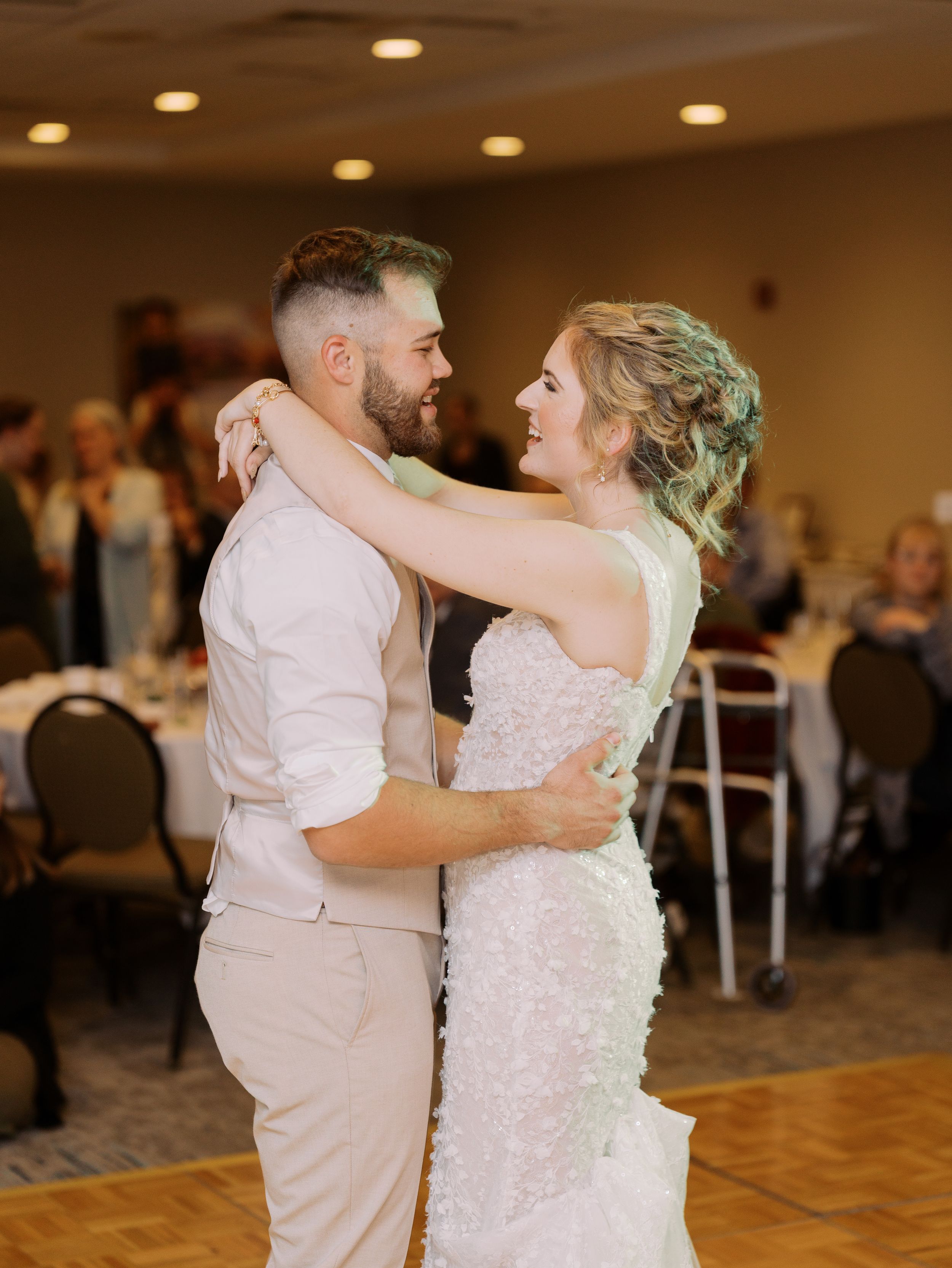 A couple shares their first dance as newlyweds in a reception hall with warm lighting and guests in the background.