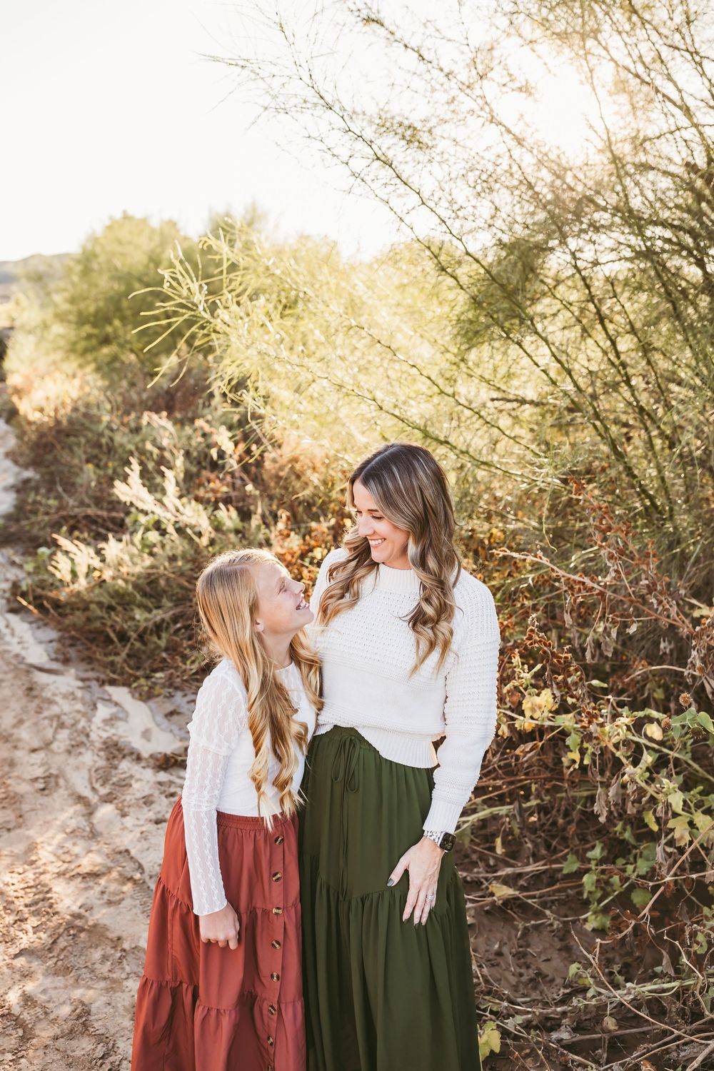 Two figures in vintage-style dresses embrace while standing against a backdrop of desert brush at sunset.