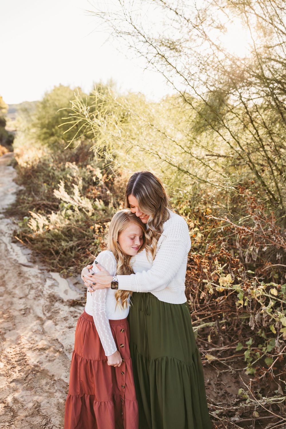 Two people in long dresses share a tender moment outdoors during golden hour with natural desert scenery.