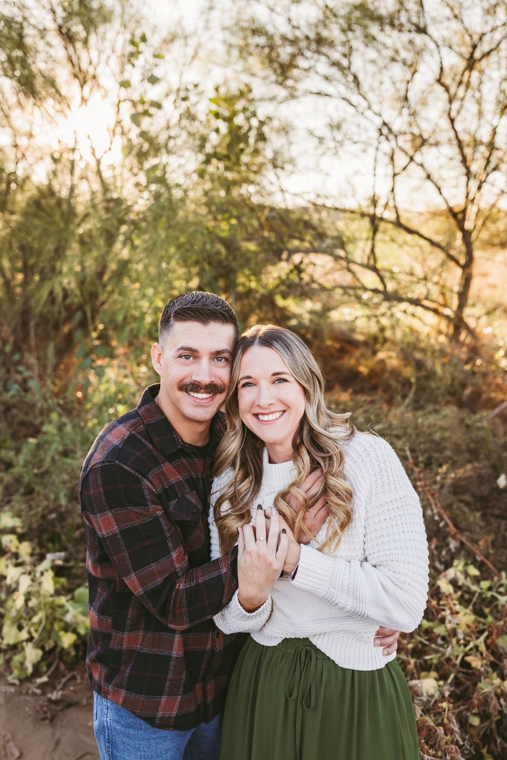 A couple poses together outdoors during golden hour, wearing casual fall attire and showing off an engagement ring.