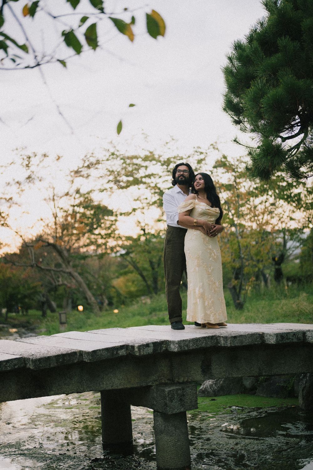 Couple embracing on a stone bridge in a Japanese garden at sunset, surrounded by lush greenery.