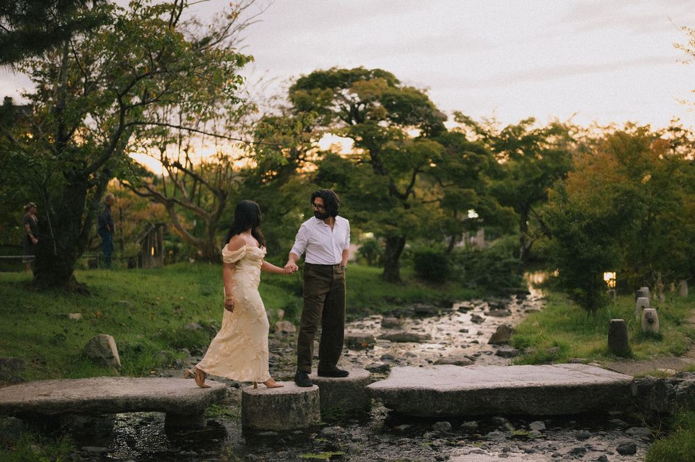 Couple holding hands on stepping stones by a stream in a lush green park at sunset.