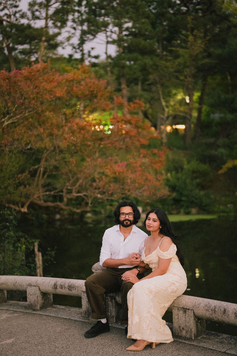 Couple sitting on a stone bridge railing in a Japanese garden at dusk with colorful autumn foliage.