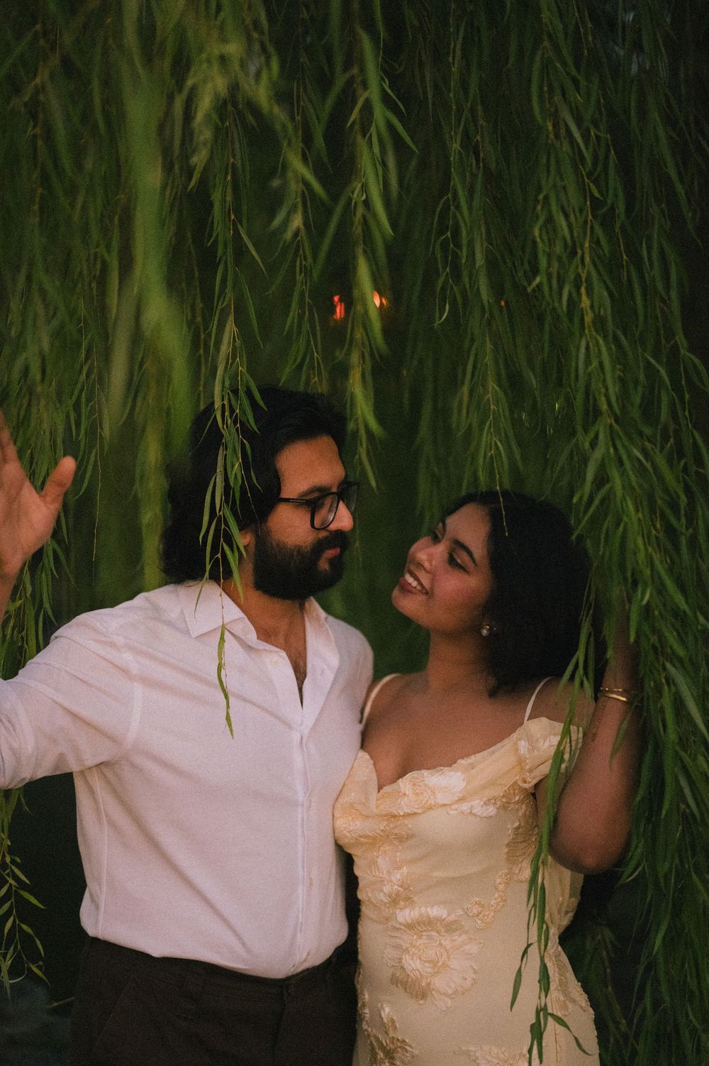 Couple smiling at each other beneath hanging willow tree branches in soft evening light.