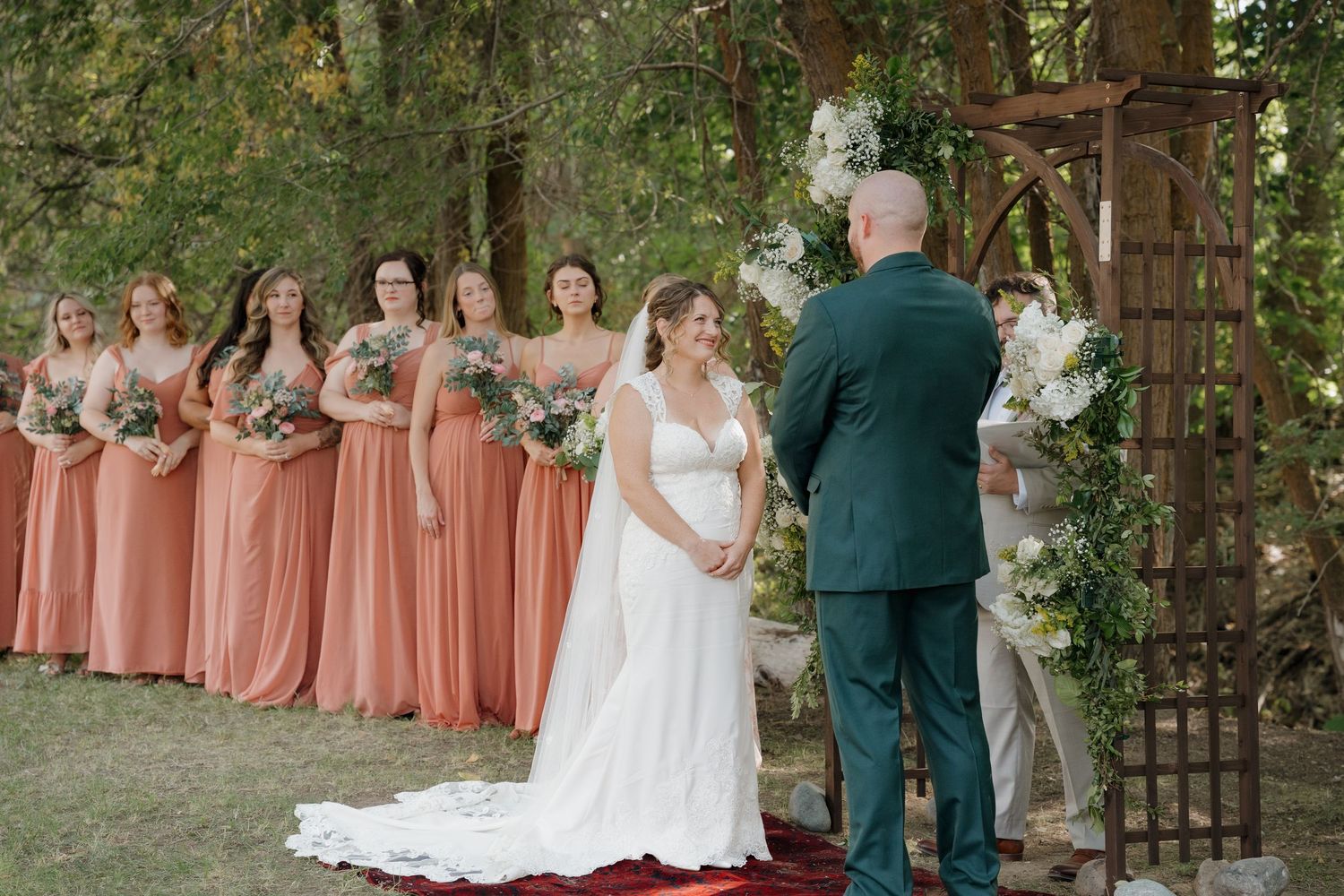 A wedding party in coral dresses and dark suits poses outdoors among trees and wooden structures. Leavenworth, WA