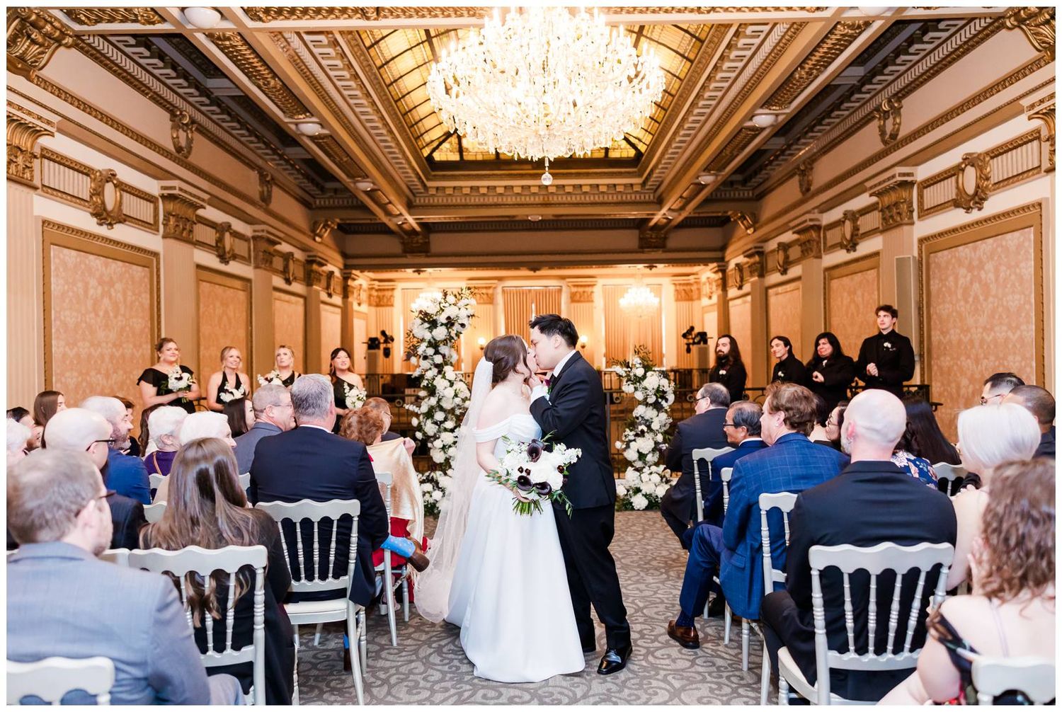 Wedding guests seated in white chairs during an indoor ceremony with crystal chandeliers overhead.