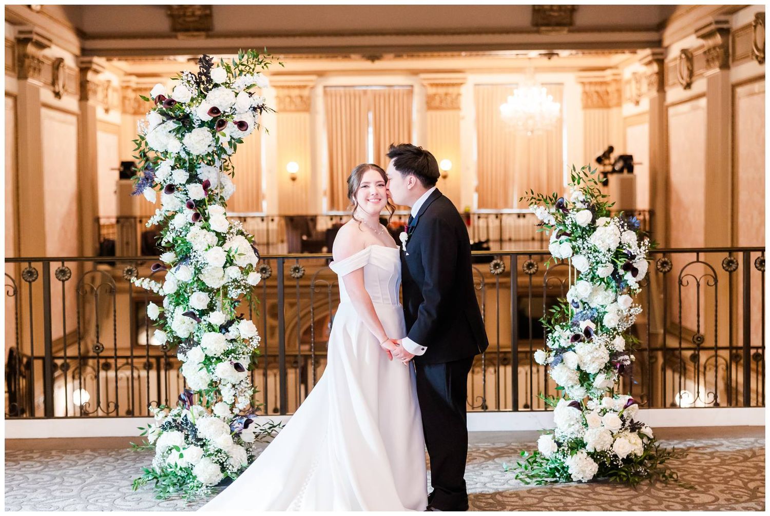 Romantic wedding ceremony setup with white floral arches and gold decor in a luxurious indoor venue.