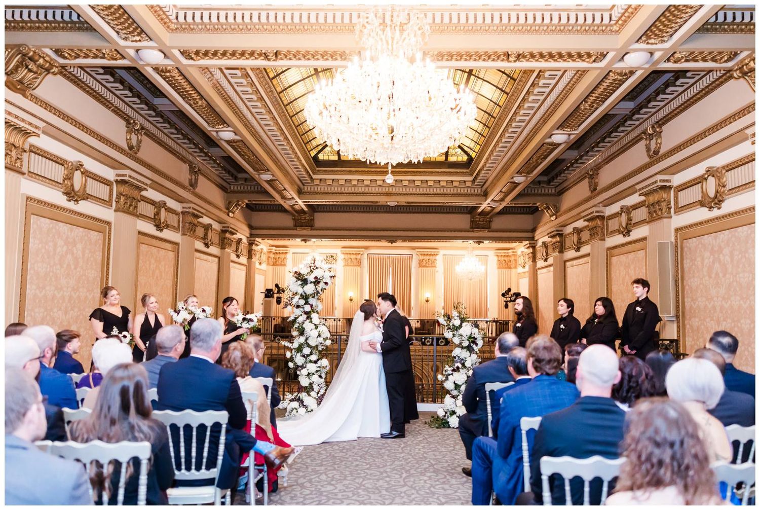 Elegant wedding ceremony in a grand ballroom with ornate chandeliers and white floral arrangements.
