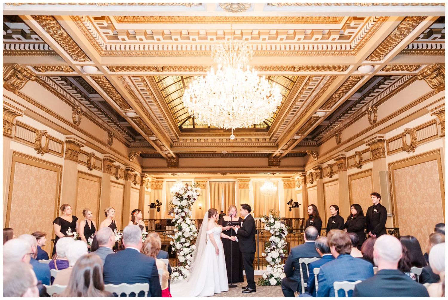 Elegant wedding ceremony in a grand ballroom with ornate ceiling details and crystal chandeliers.