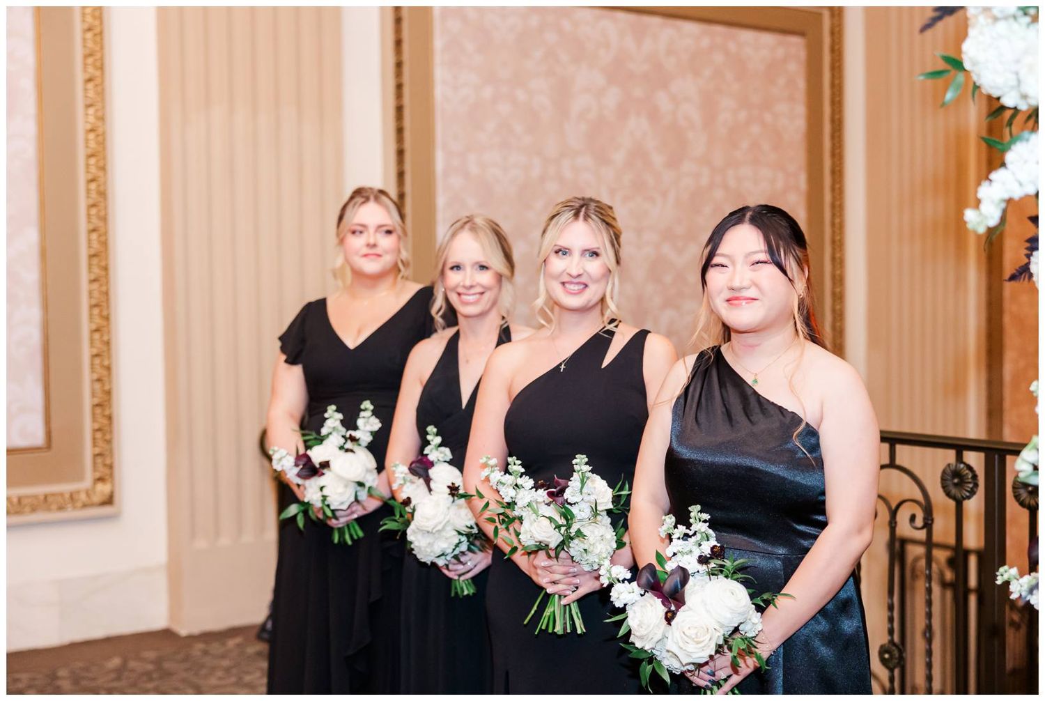 Wedding party members in black dresses holding white floral bouquets pose in an elegant ballroom setting.