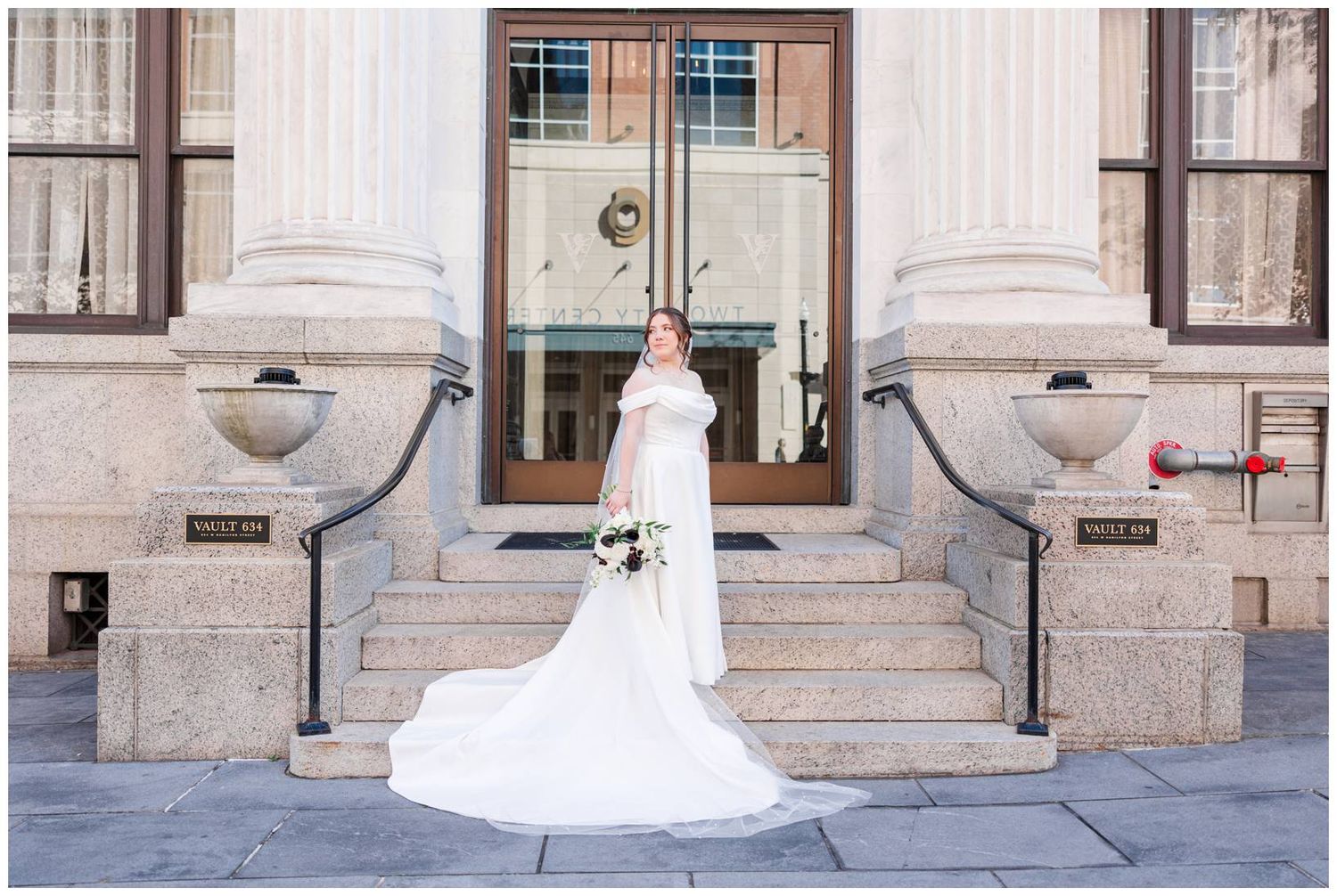 Bride in flowing white gown poses on stone steps of classic building entrance.
