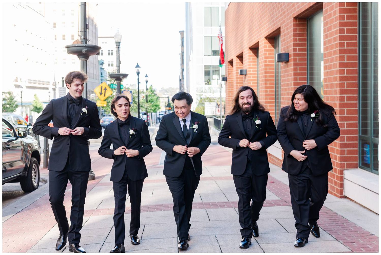 Group wearing formal black attire walking together along a brick-lined sidewalk in an urban setting.