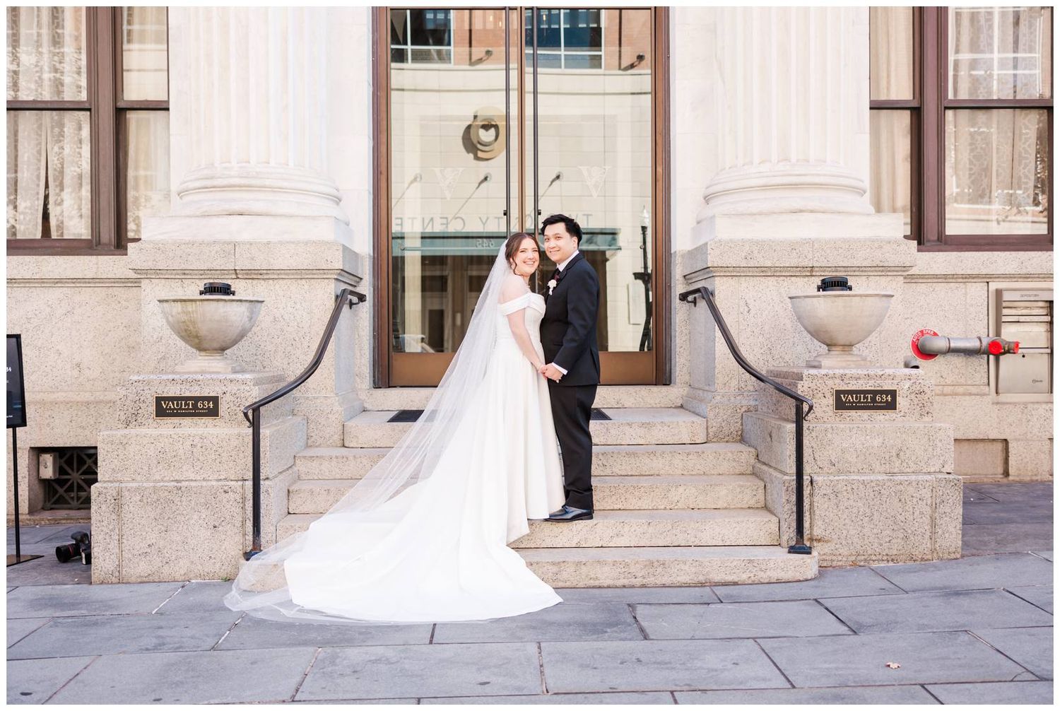 Newlyweds pose on grand stone steps of historic building with bride's dress trailing behind.