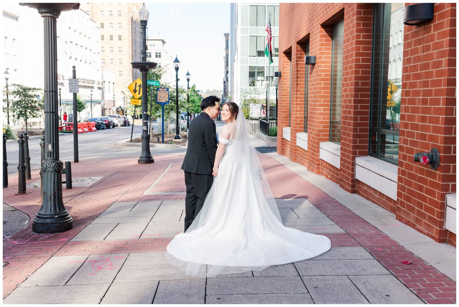 Wedding couple poses in front of brick building with flowing veil creating romantic urban portraits.