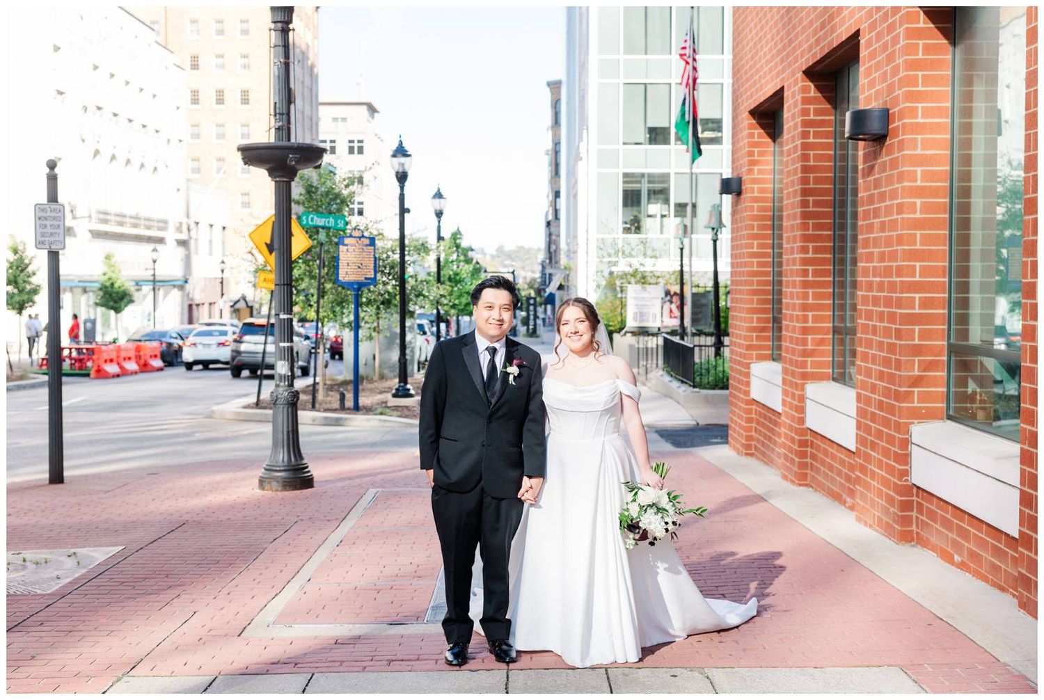 Wedding couple walks together on brick sidewalk past historic red brick buildings in downtown area.
