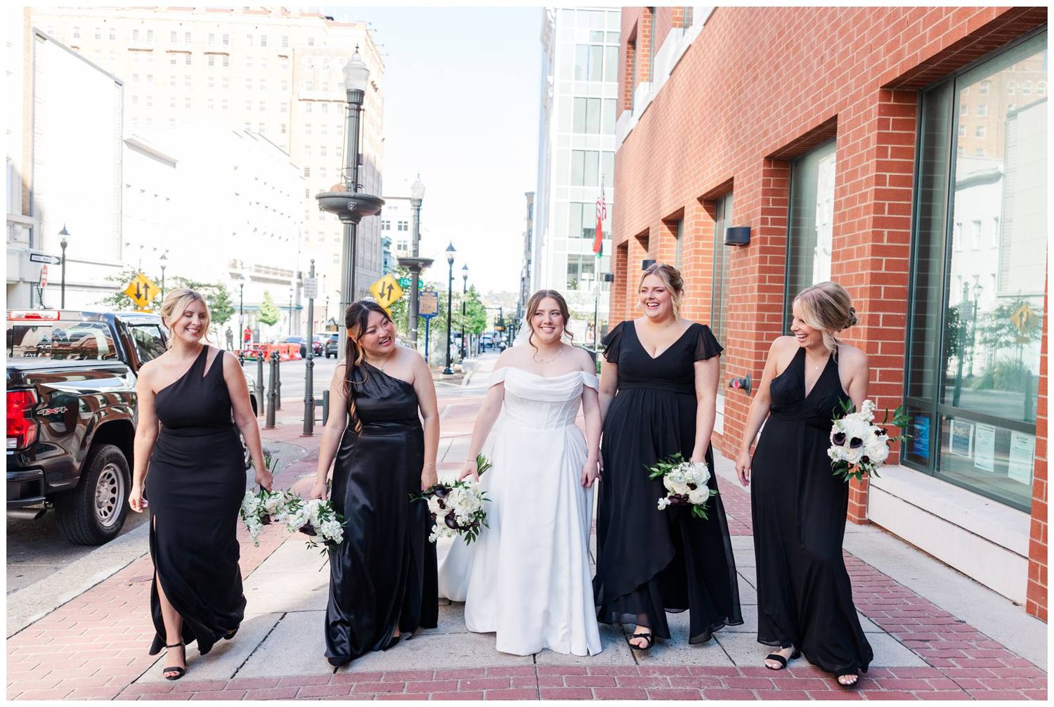 Wedding party poses on city sidewalk with bride in white gown and bridesmaids in black dresses.