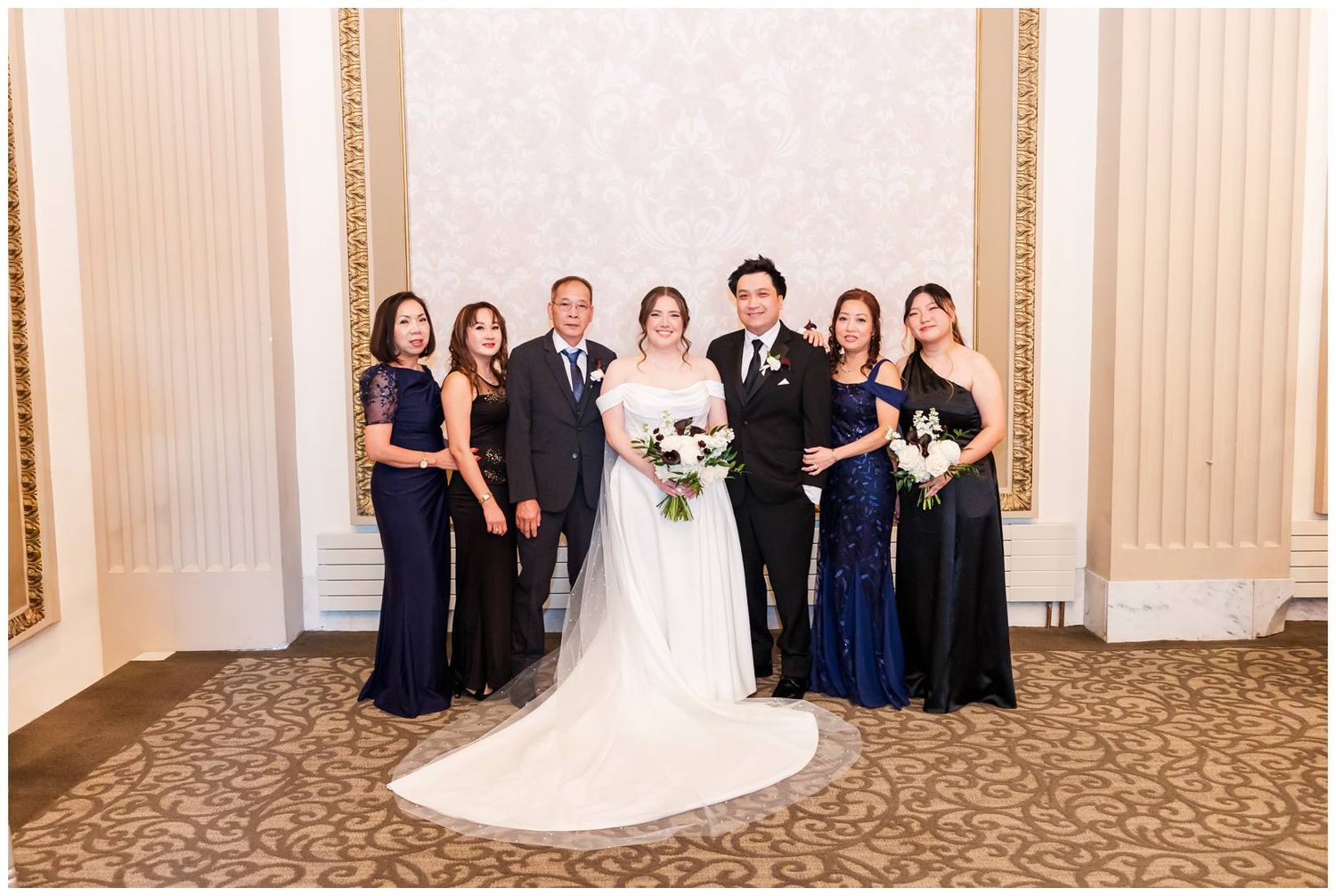 Wedding party poses in formal attire against a cream-colored wall with decorative molding.