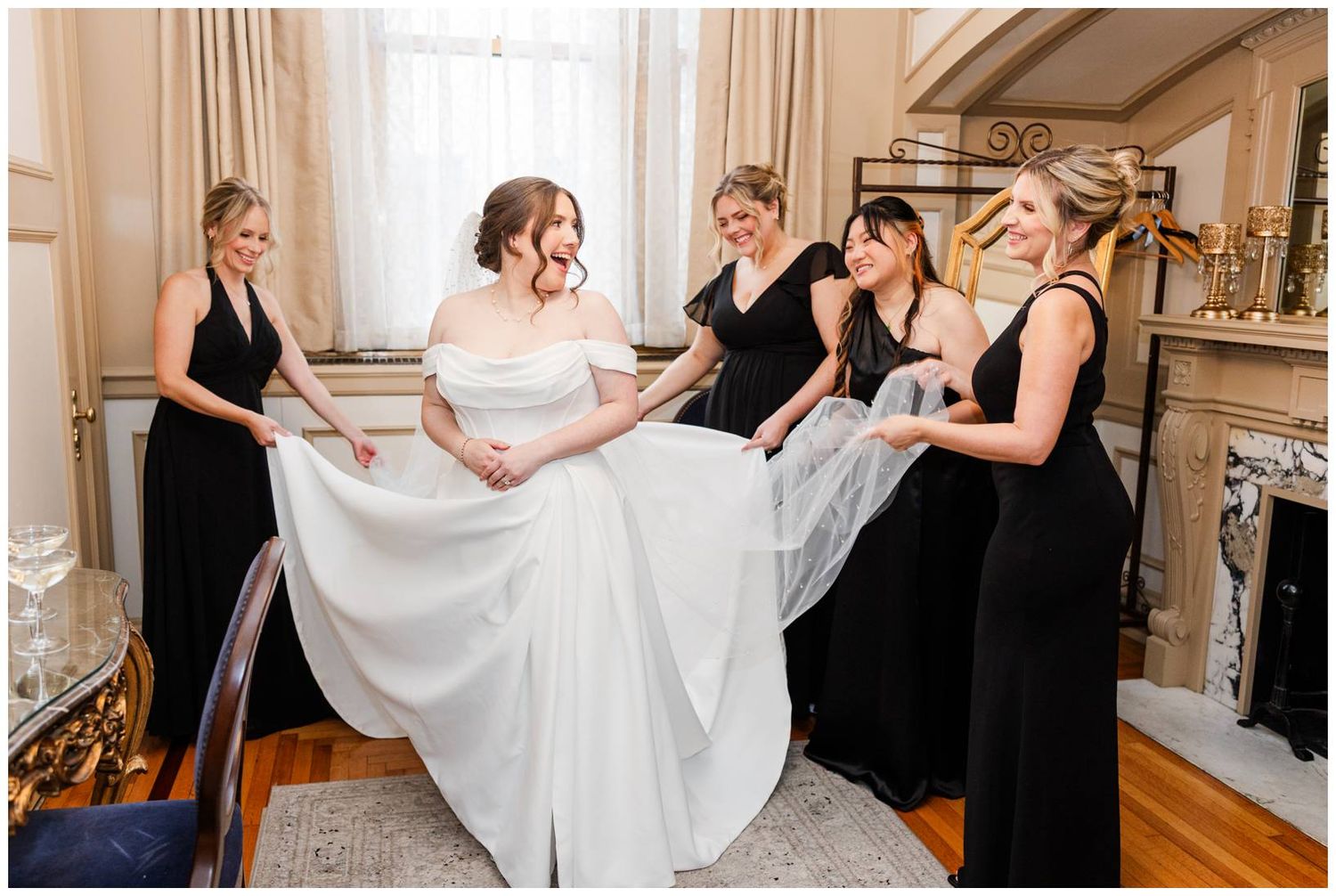 Bridesmaids in elegant black dresses help adjust the bride's flowing white gown in an ornate room.