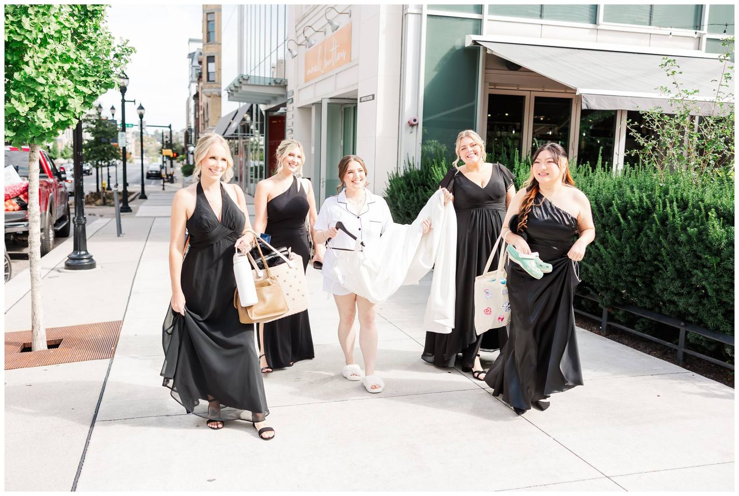 Wedding party walking down a city sidewalk with bridesmaids in black dresses and bride in white.