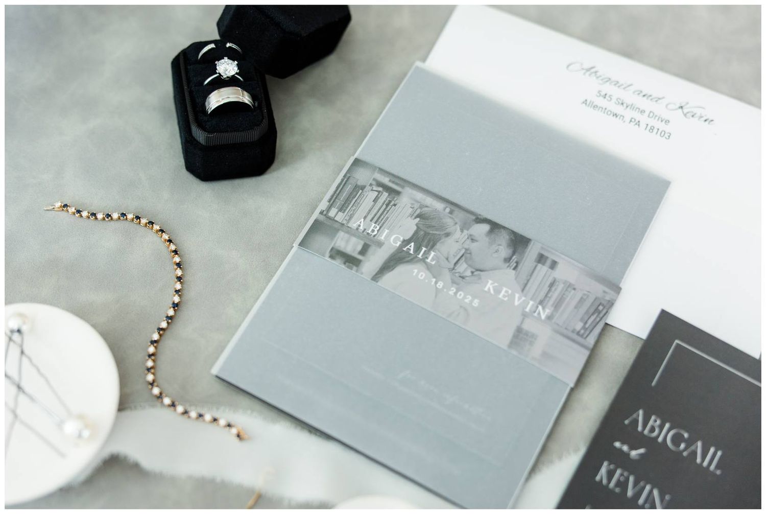 A wedding invitation suite with a black ring box and delicate rose gold beaded details on white marble surface.