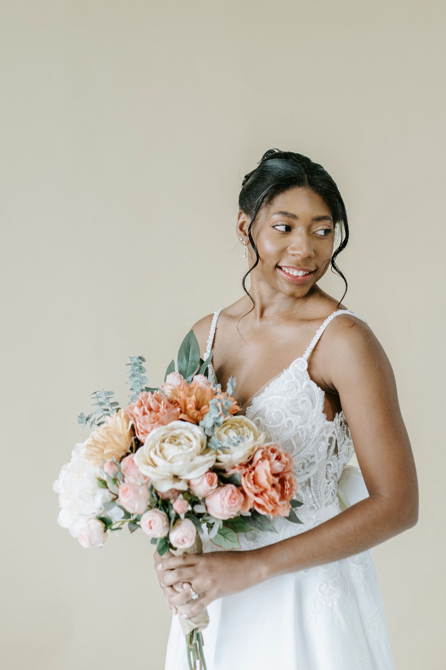 A bride in a white lace dress holds a romantic bouquet of pink and white roses against a neutral background.