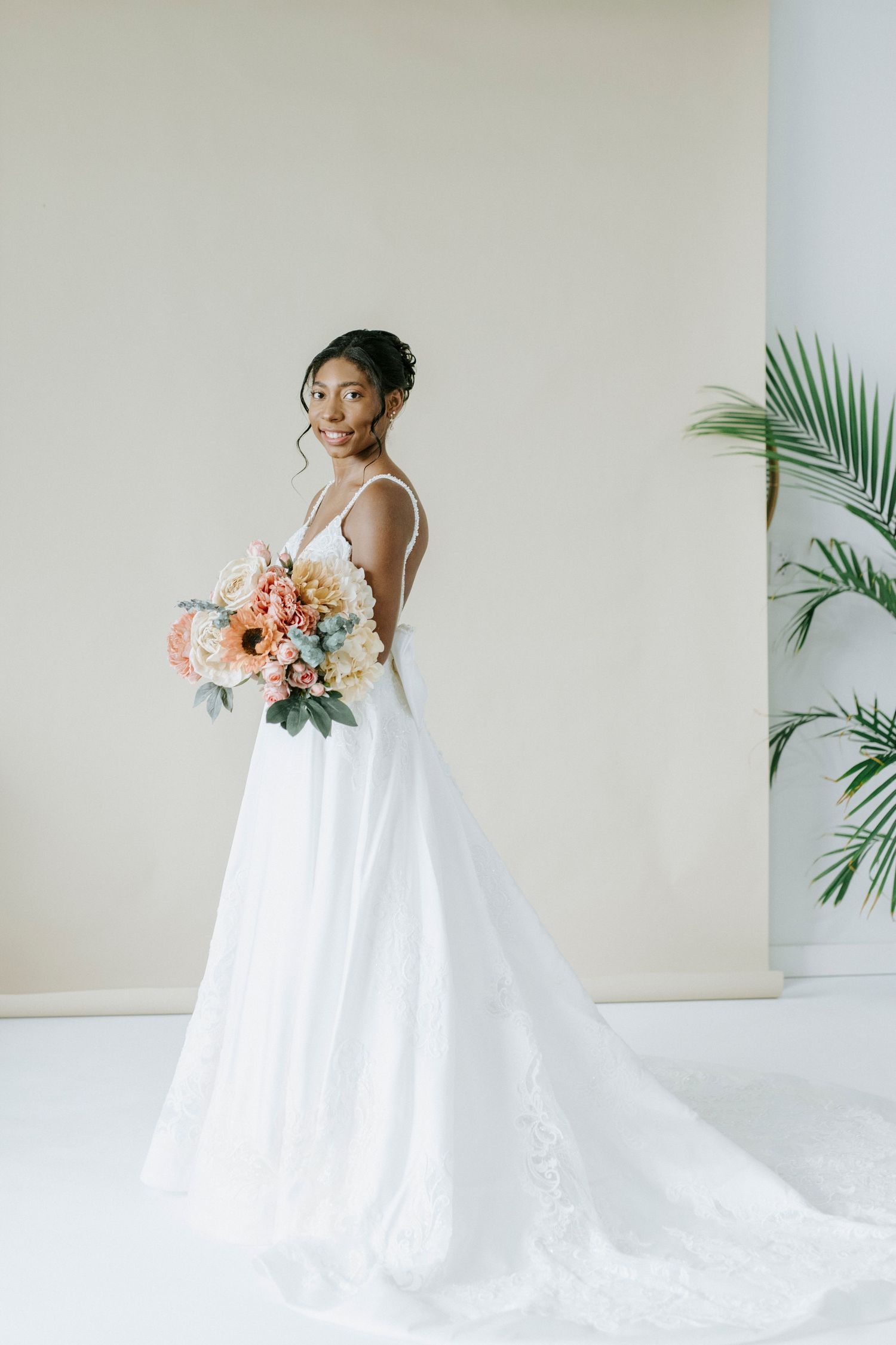 Elegant bride in flowing white ballgown poses with pink and peach bouquet against minimalist backdrop with tropical palm leaves.