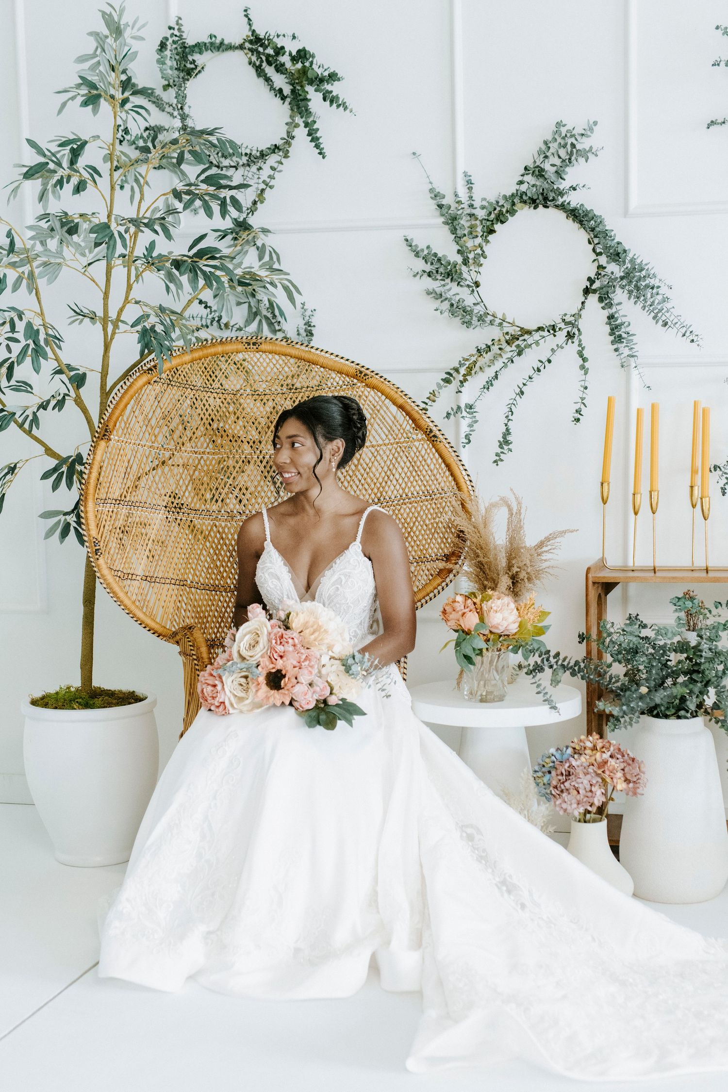 Elegant bridal portrait with peacock chair, white gown, pink bouquet and botanical decor against white backdrop.