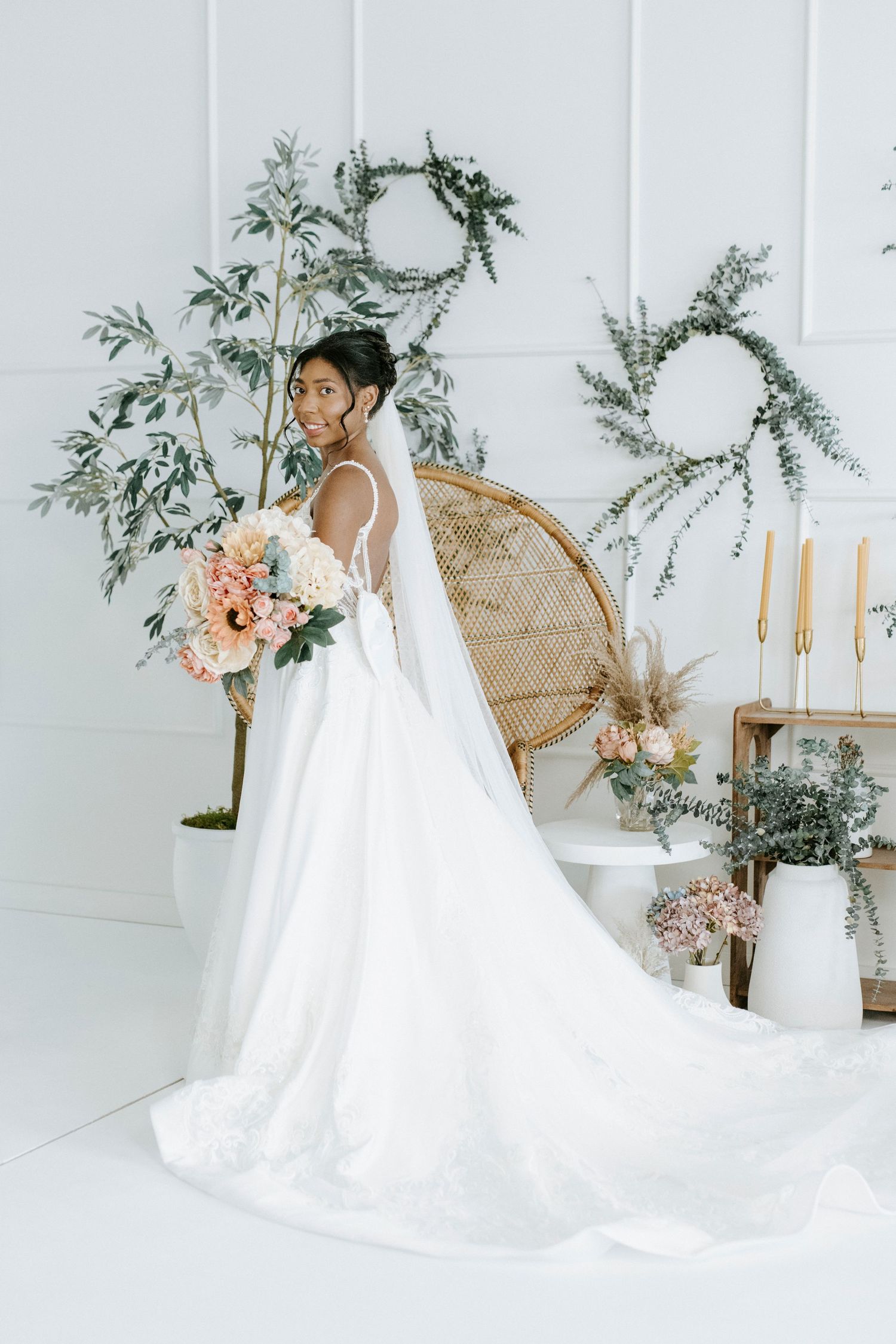 A flowing white wedding dress with a long train displayed in a bright studio setting with floral accents and rattan furniture.